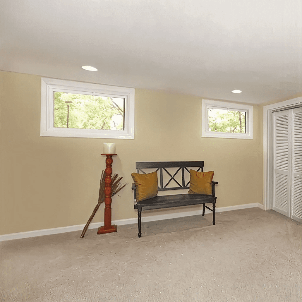 Interior view of a finished basement living area featuring two high-set white hopper windows, a dark wooden bench with mustard pillows, a tall candle holder, and beige carpet.