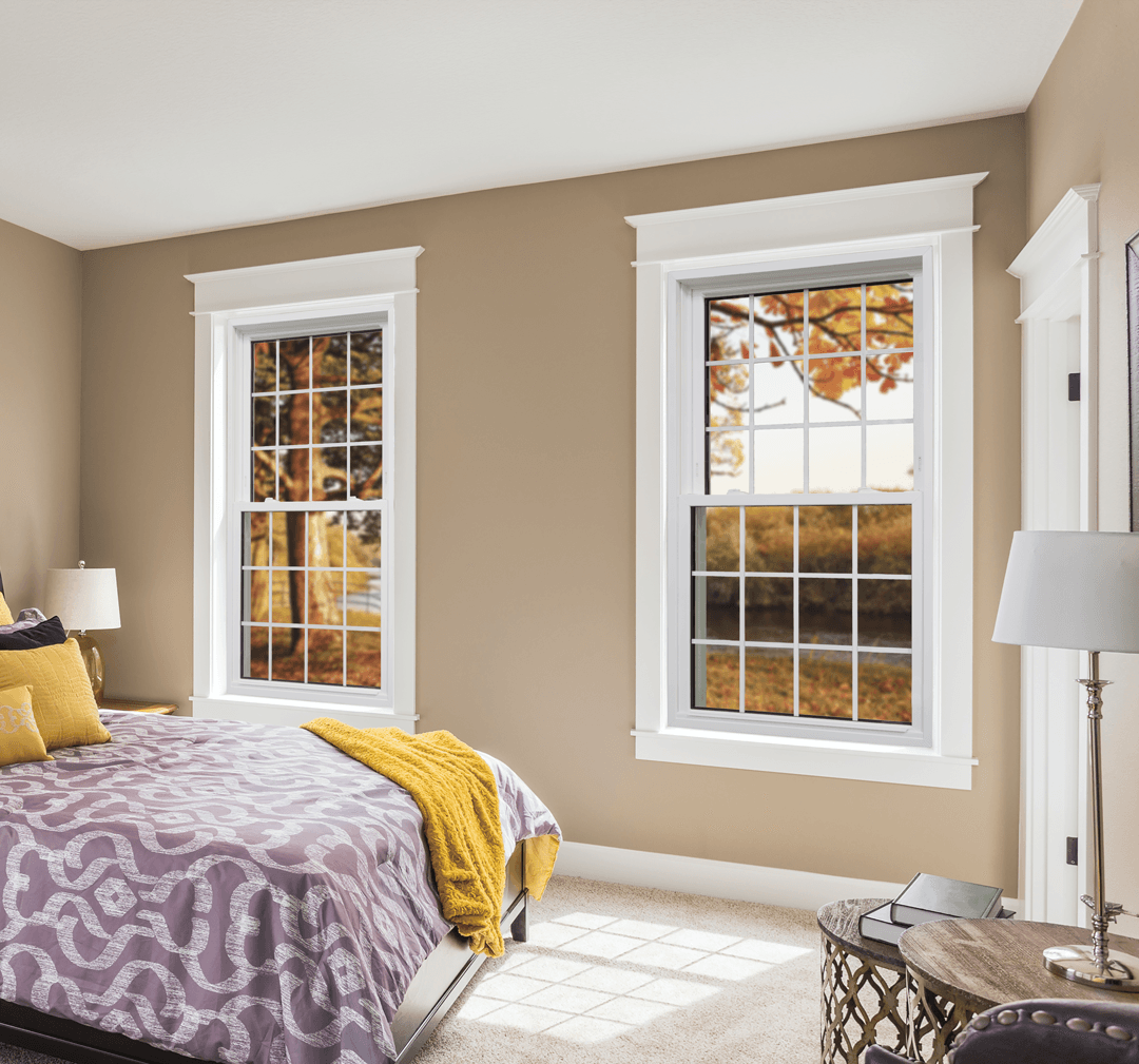 Interior view of a bedroom featuring two white double-hung windows with colonial grilles, looking out onto trees with golden autumn leaves.