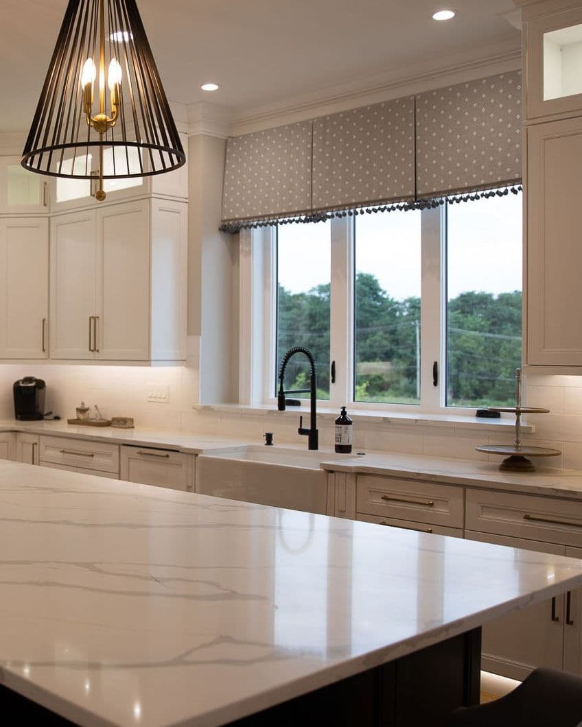Modern kitchen featuring a large marble island, white cabinetry with brass hardware, an apron-front sink with a black faucet set below a three-pane casement window with a patterned grey valance, and a cage pendant light hanging overhead.