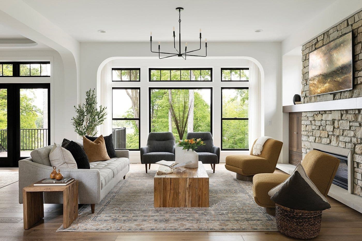 Interior view of a modern living room featuring three large, black-framed picture windows with upper transoms set within a decorative wall arch, complemented by a grey sofa, mustard accent chairs, a stone fireplace, and a light wood coffee table.