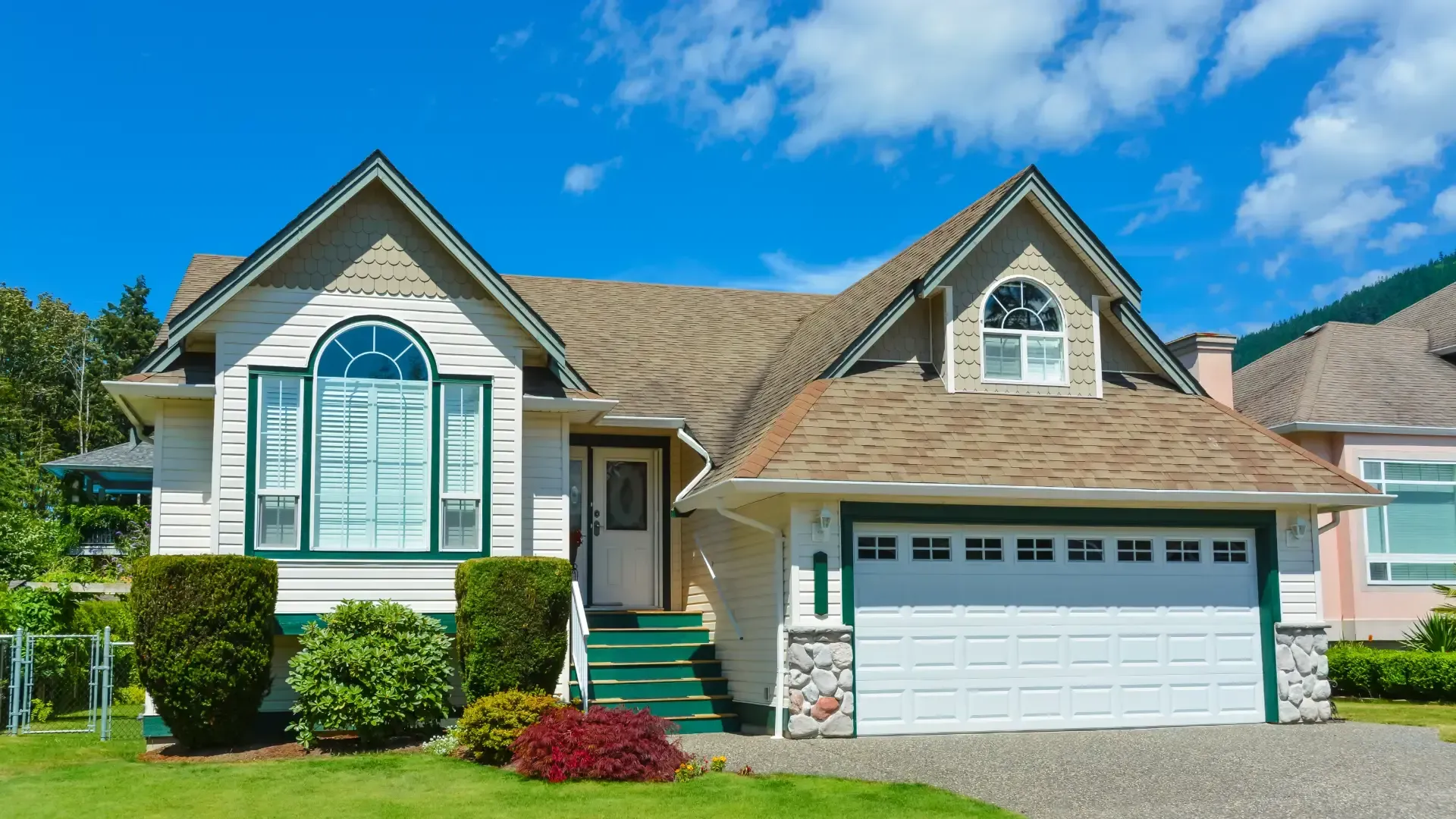 Suburban home featuring cream siding, green trim around arched accent windows, a front garage, and green porch steps under a bright blue sky.