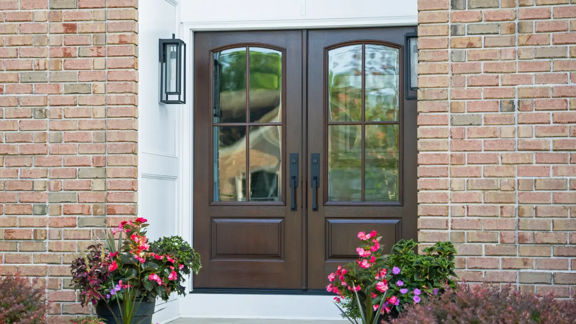 Exterior view of dark hazelnut double French doors featuring four-lite arched glass inserts, matte black vertical handles, and black lantern sconces against light brick and white casing.