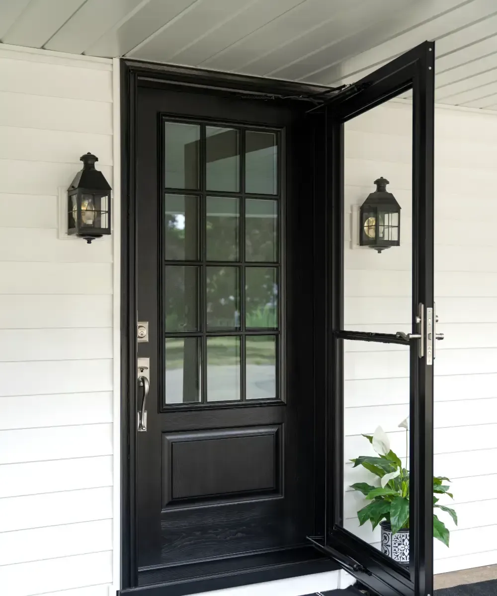 Exterior view of a coal black front door featuring a glass inset with colonial grilles and satin nickel hardware, set behind an open matching black glass storm door against white lap siding with flanking lantern wall sconces.