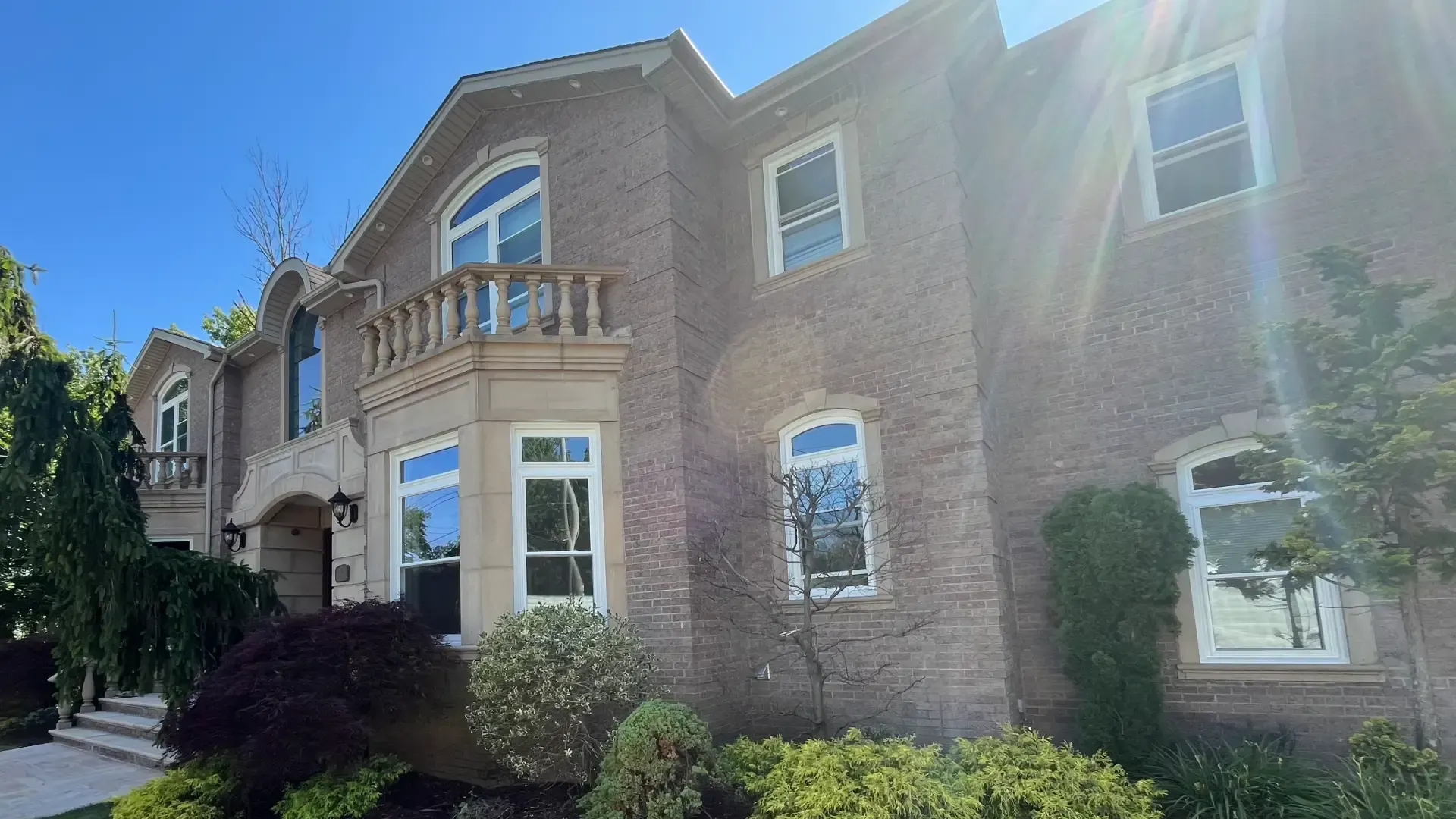 Exterior view of a large two-story stone mansion with white-framed double-hung and arched windows under bright sunlight, accented by a second-story stone balustrade balcony and lush landscaping.