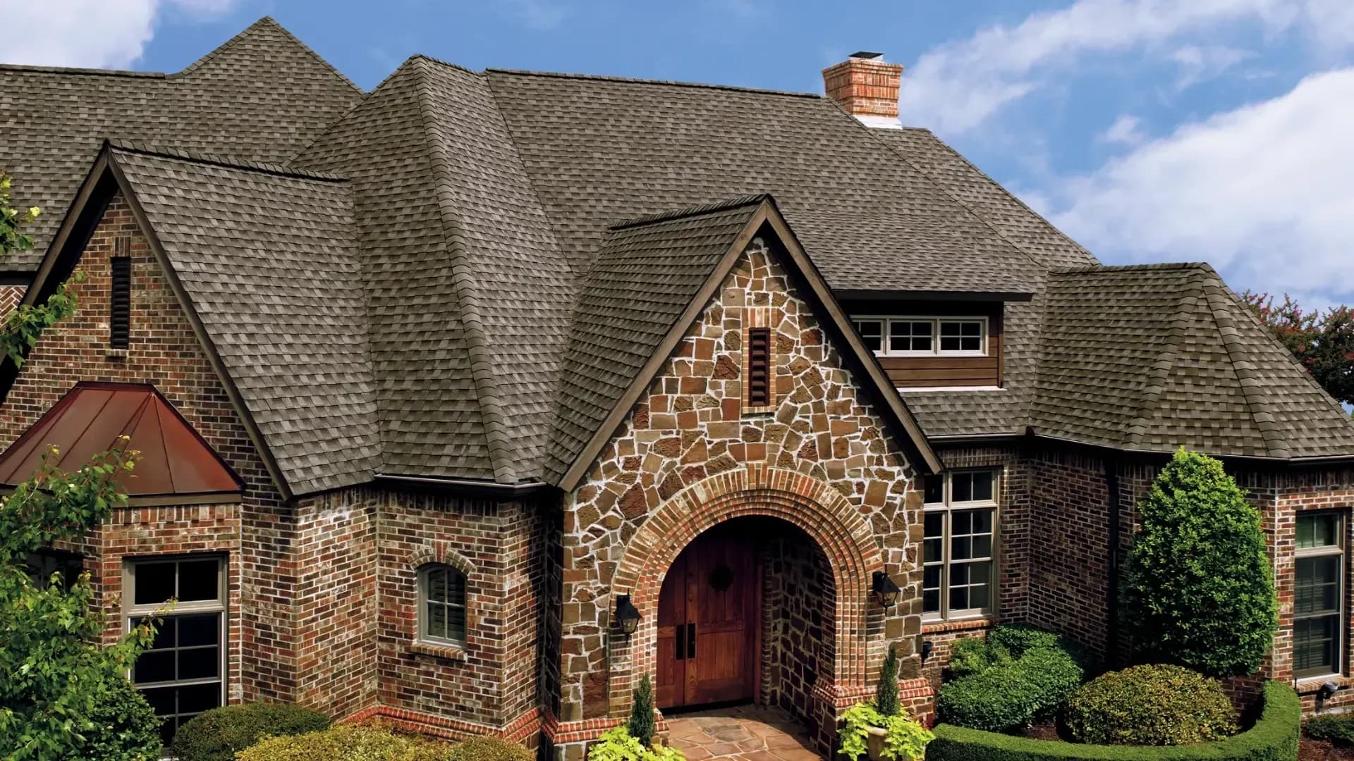 Exterior front view of a Tudor-style home featuring Weathered Wood architectural asphalt shingles on a multi-gabled roof, framed by a rustic fieldstone arched entryway, red brick walls, copper bay window accents, and manicured landscaping.