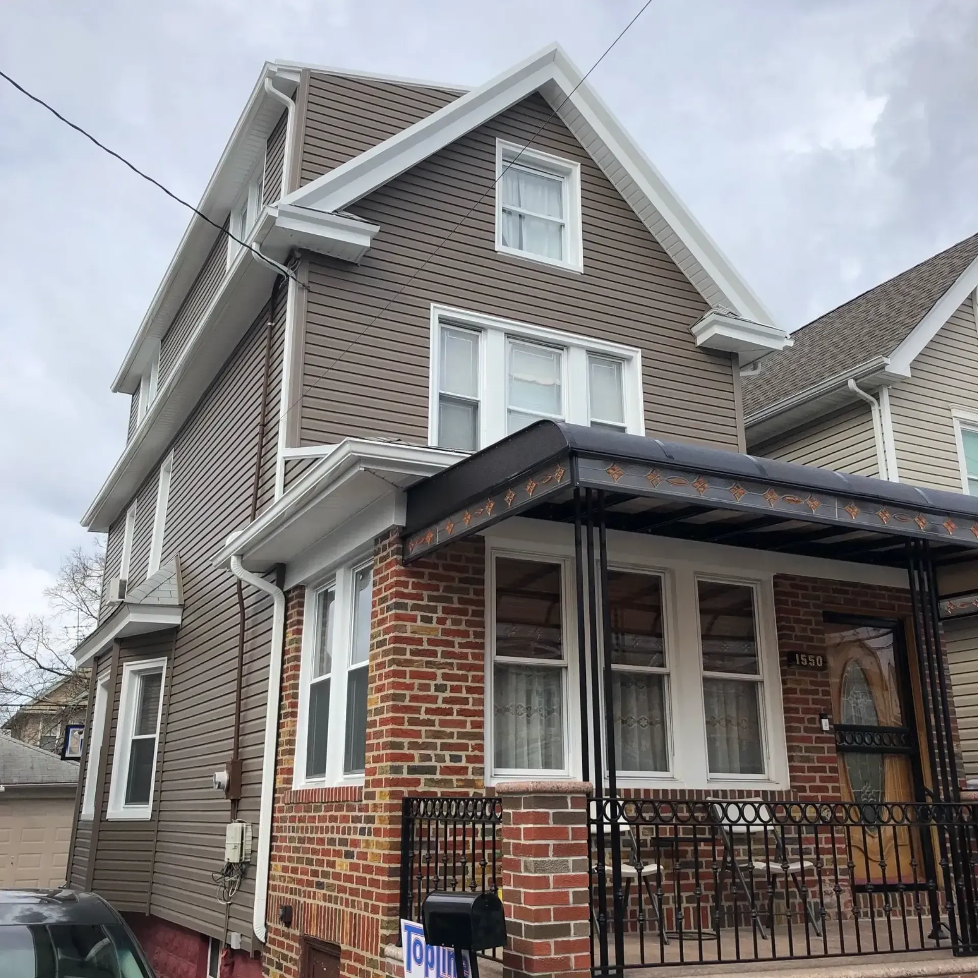 Exterior perspective view of a multi-story home featuring taupe horizontal siding, white trim, brick base construction, and a covered front porch with black iron railings.