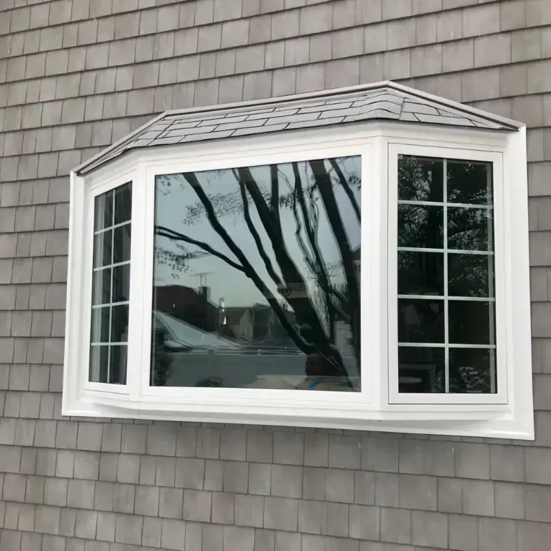 White vinyl bay window with a shingled mini-roof, featuring a large center picture window and gridded side windows, set into grey shingle siding.