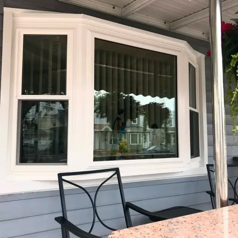 White vinyl bay window with a central picture pane and flanker double-hung windows, installed under a covered porch with dark outdoor chairs in the foreground.