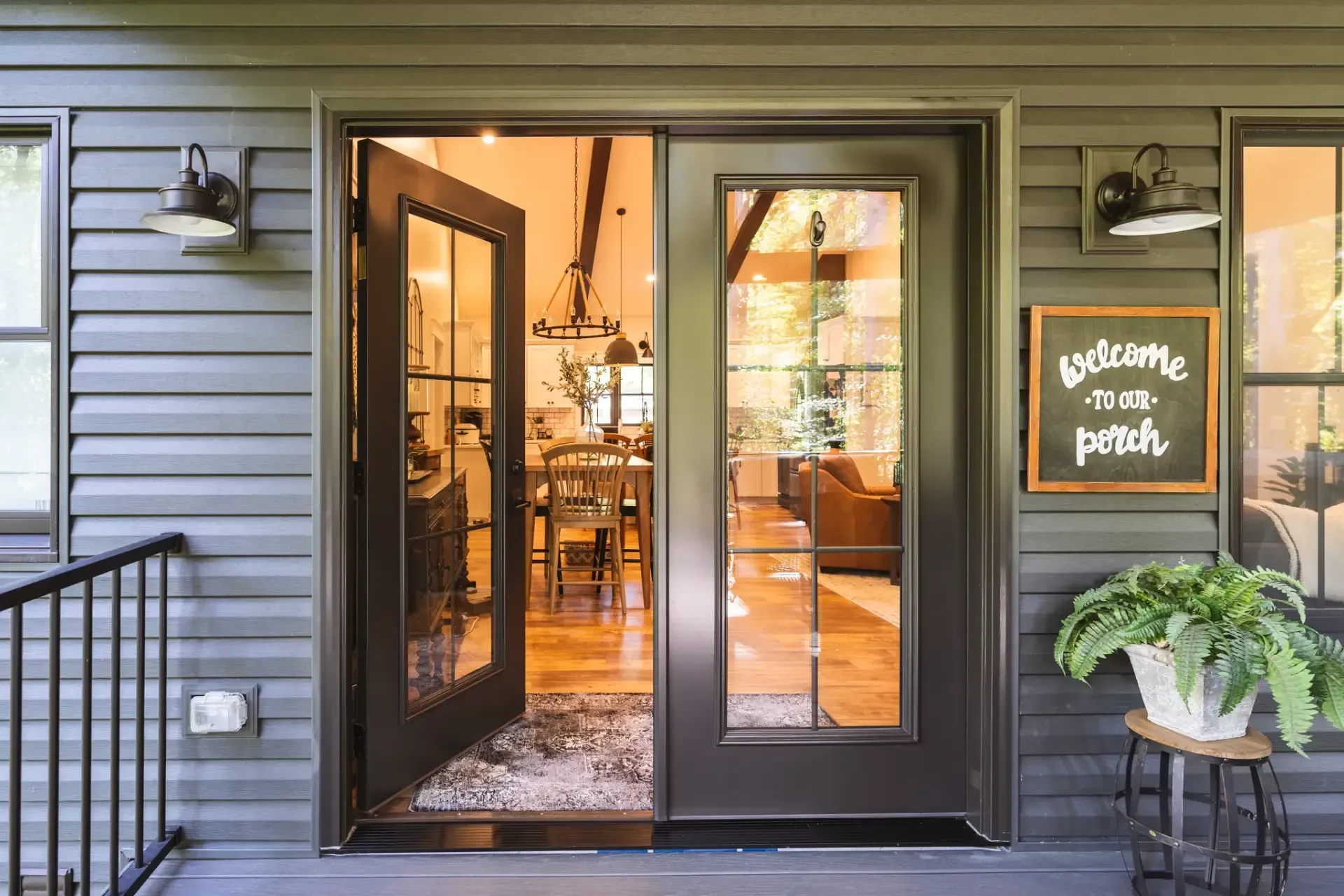 Exterior view of open dark bronze steel French doors with full-length glass panes and internal grilles, connecting a covered porch to a warmly lit home interior.