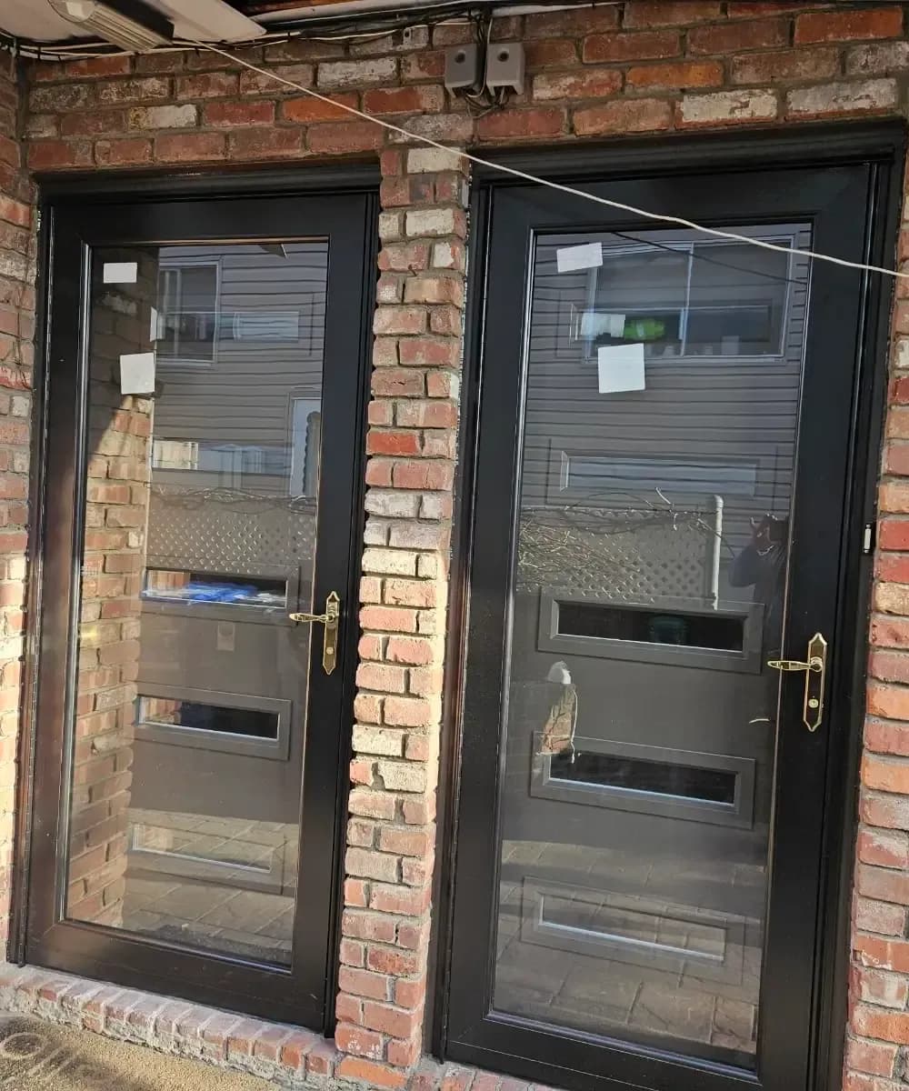 Exterior view of two side-by-side black storm doors with full glass panes and brass handles, installed over grey entry doors with horizontal glass cutouts within a brick facade.