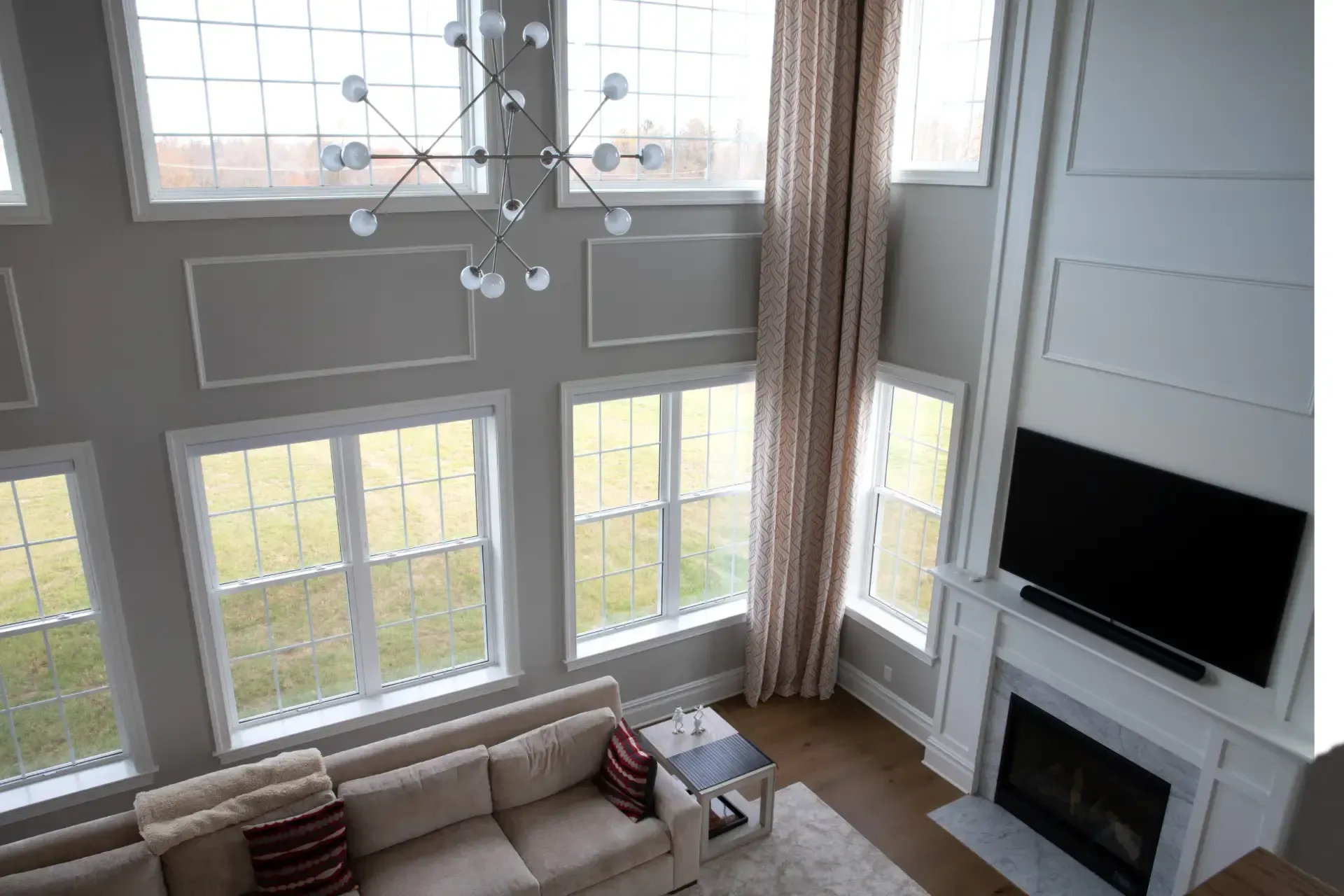 High-angle interior view looking down into a modern two-story great room, featuring a two-story wall of white-framed multi-pane windows, a sputnik chandelier, beige sectional sofa with accent pillows, floor-to-ceiling drapes, and a fireplace mantle with a mounted television.