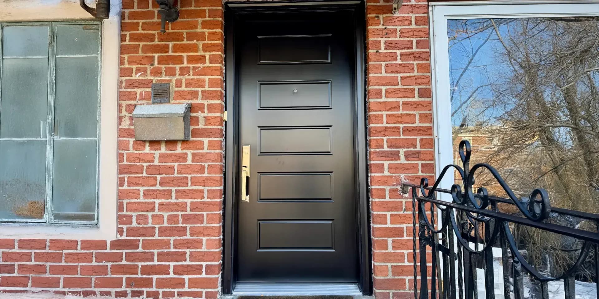 Exterior view of a modern matte black front door with five horizontal raised panels and gold/brass handleset hardware, installed between red brick walls with a frosted glass window on the left and a wrought-iron porch railing on the right.