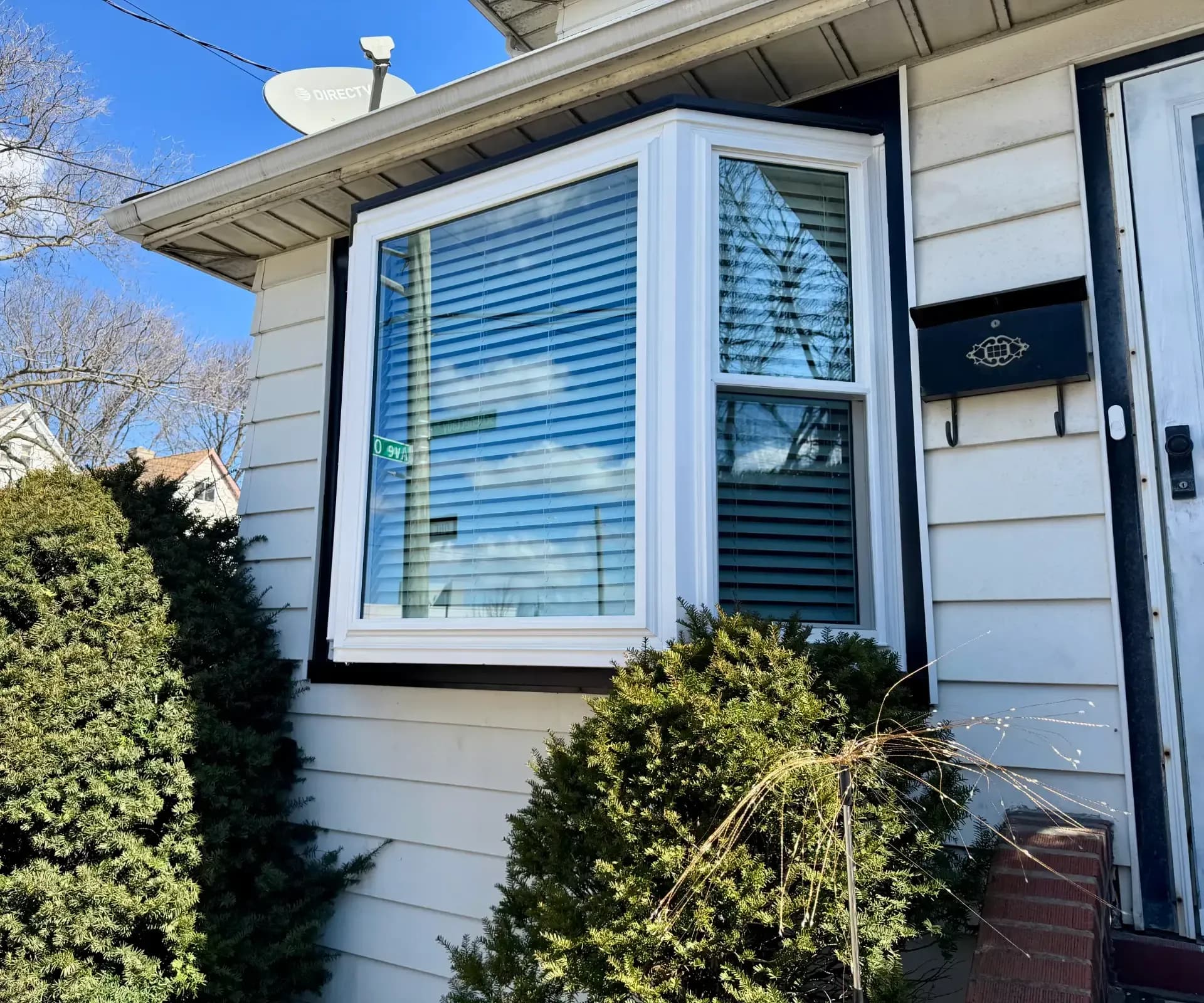 White vinyl bay window with dark trim and interior blinds, set on a house with light siding and surrounded by green shrubs.