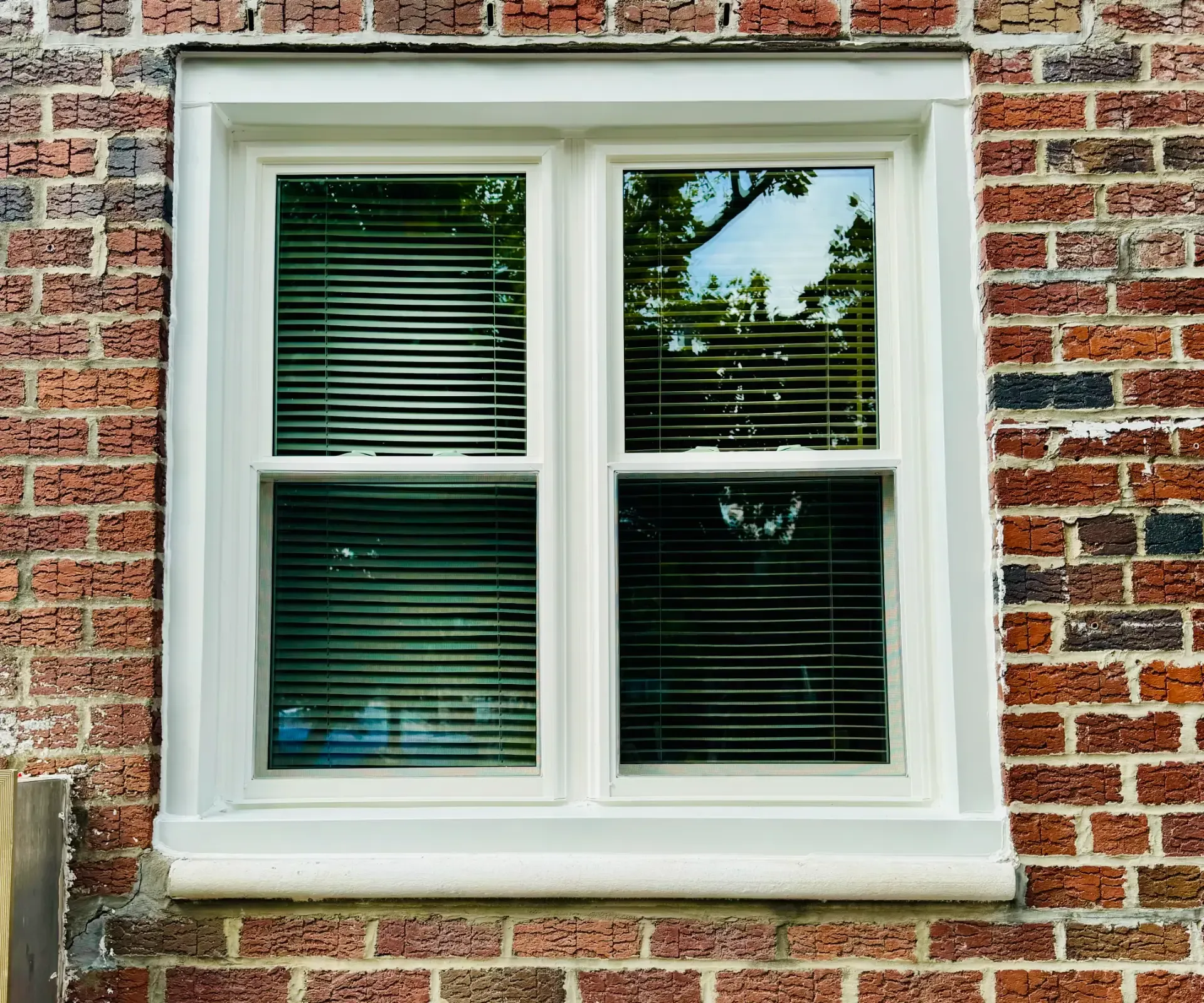 Exterior view of a white double-hung window set with horizontal blinds, installed in a red brick wall.