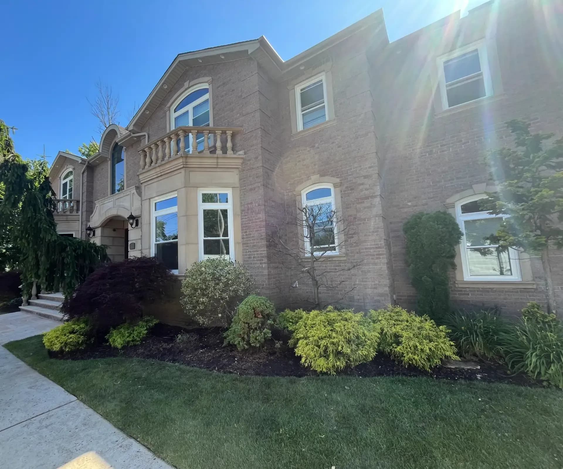 Exterior of a large two-story brick mansion with white-trimmed windows, a stone balcony, and manicured landscaping under a bright, sunny sky.
