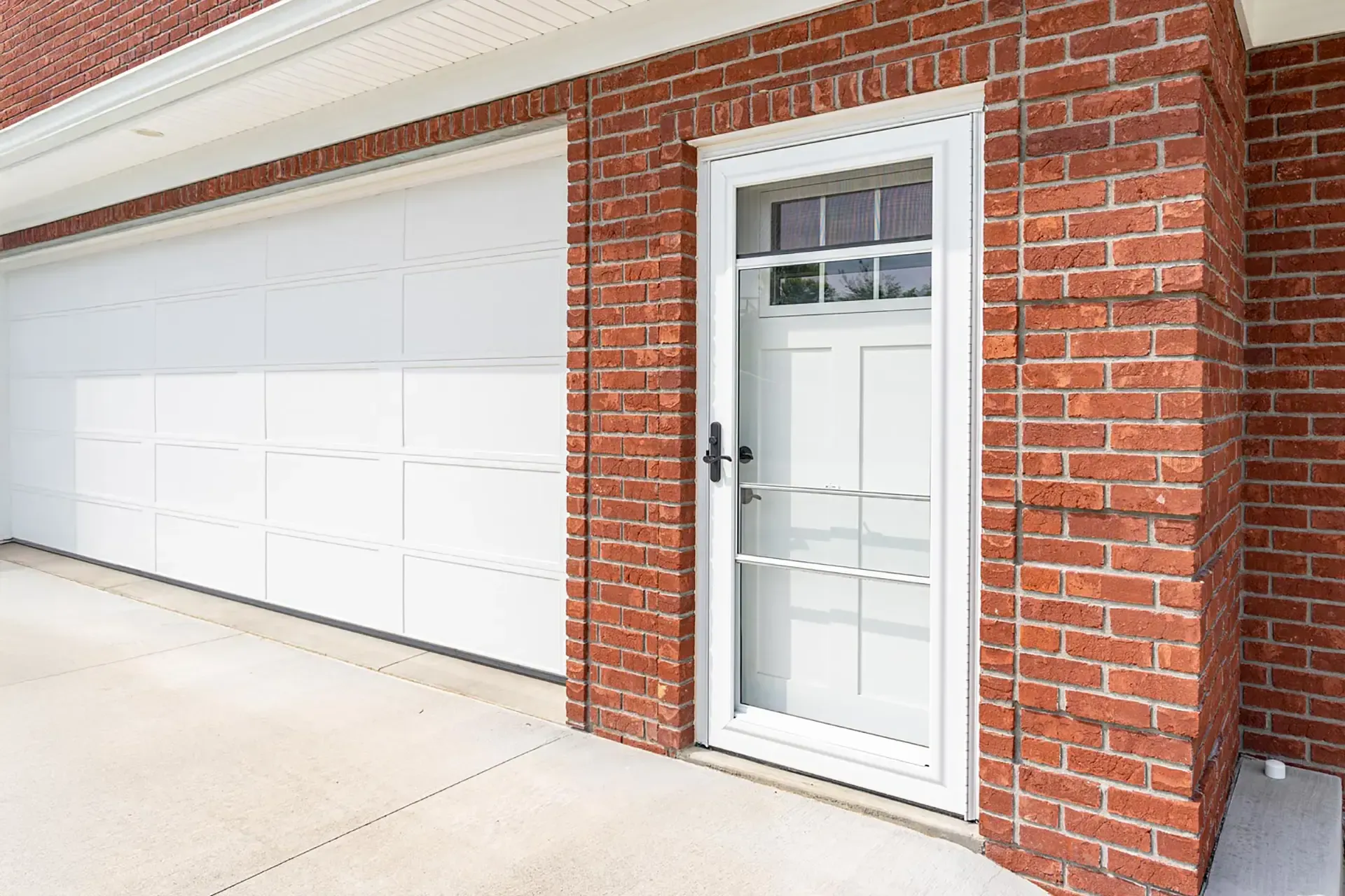 Exterior view of a white glass storm door protecting a white side door adjacent to a large garage door on a red brick home.