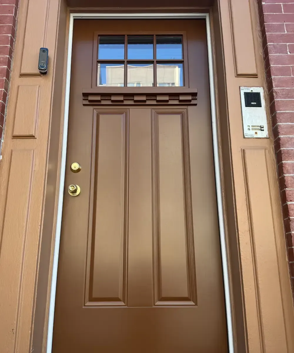 Exterior view of a dark brown Craftsman-style front door featuring a 6-pane window unit at the top, a shelf-like dentil molding ledge underneath, and two long vertical lower panels, framed by matching brown trim against a brick wall.