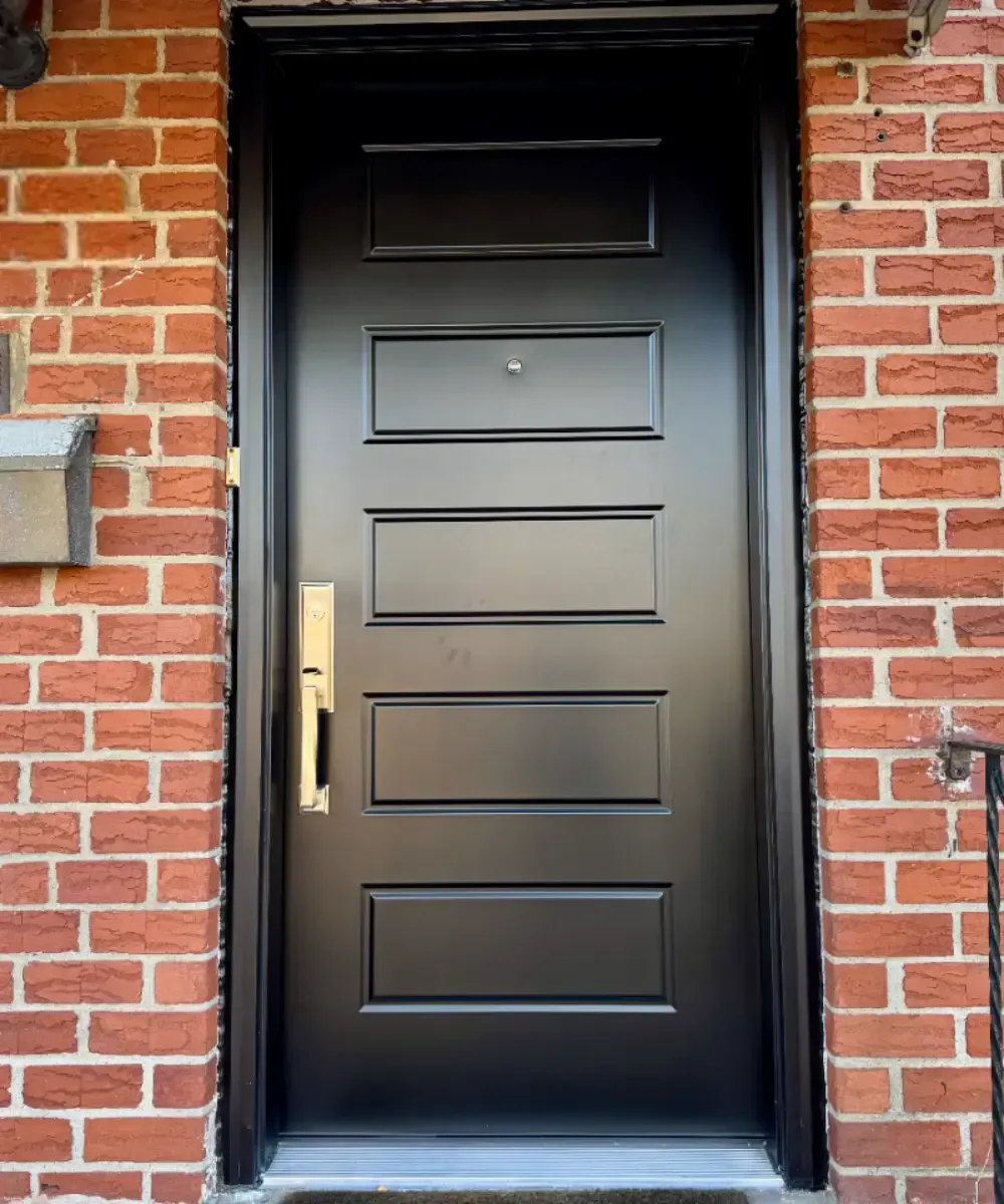 Exterior view of a sleek, matte black front door with five horizontal raised panels and modern brass handleset hardware, set within matching black frame molding against a red brick wall.