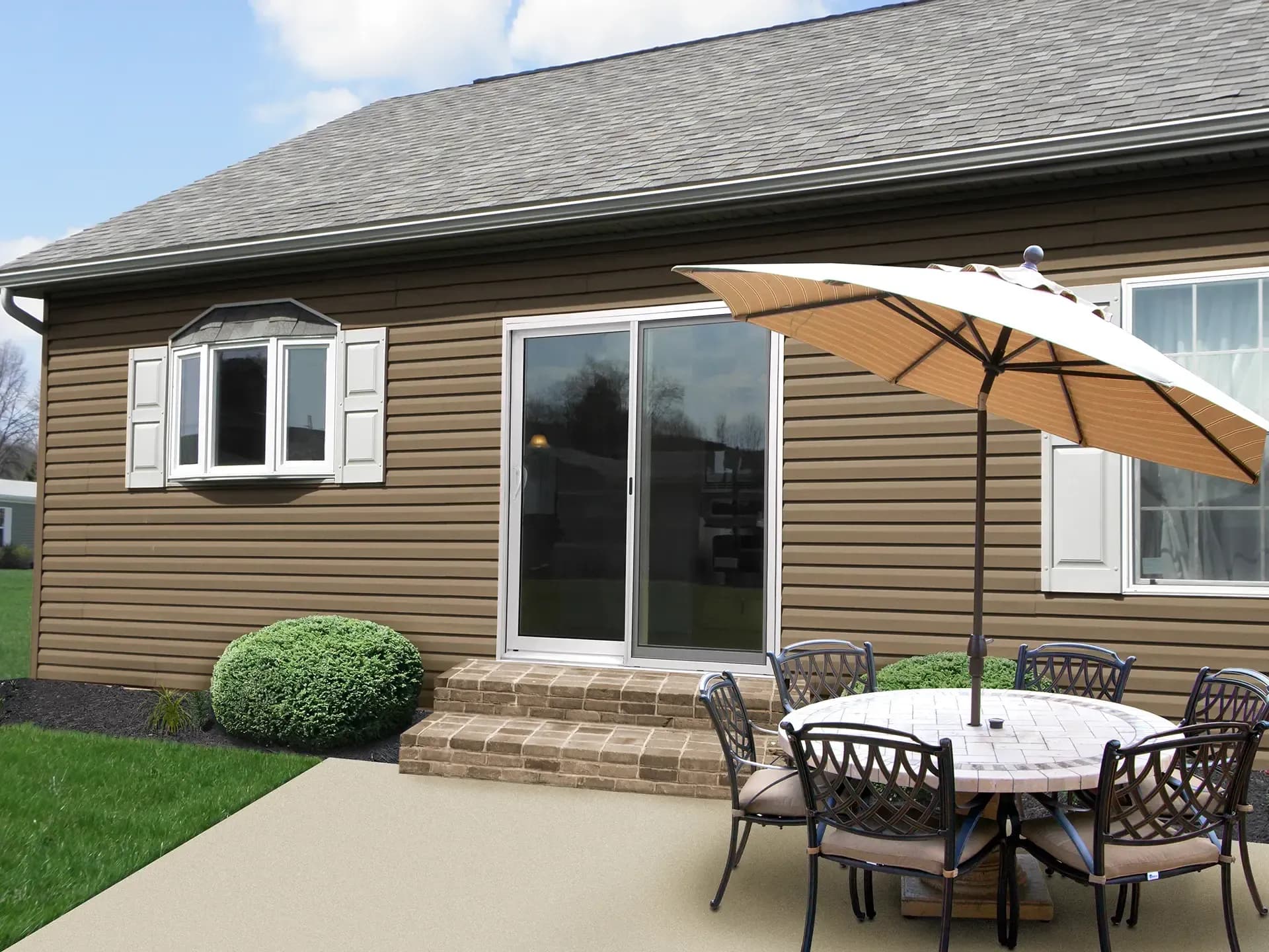 Exterior view of a backyard patio featuring brown horizontal siding, a white-framed sliding glass patio door set above brick steps, and a dining table setup with an open patio umbrella.