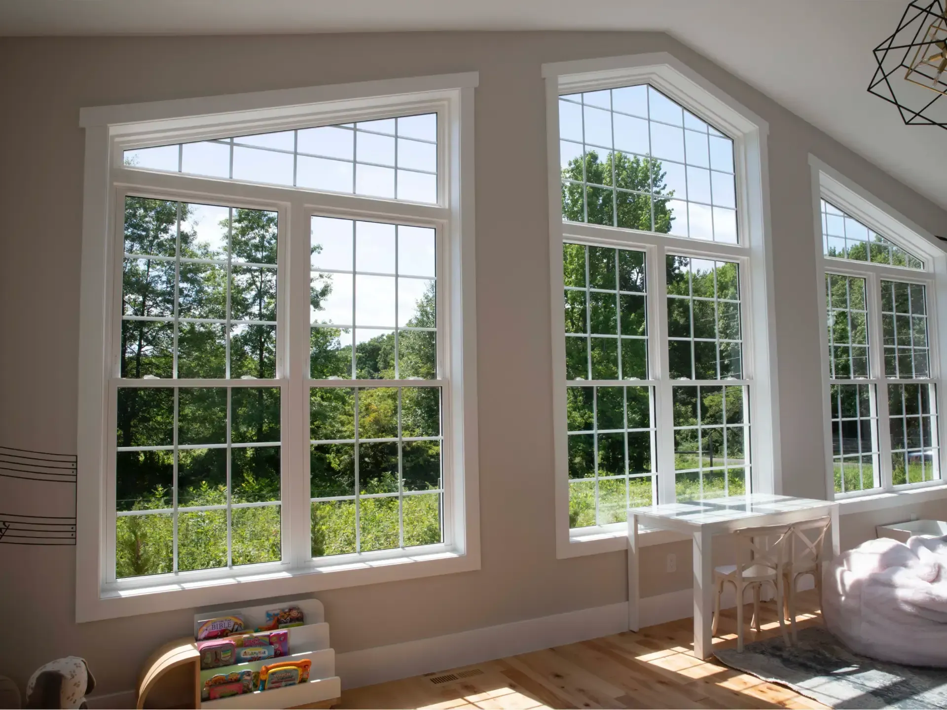 Large white-framed interior windows with colonial grilles following the vaulted ceiling line, set in a bright room featuring light hardwood floors, a children's activity table, and a plush beanbag chair overlooking a sunny, tree-lined view.