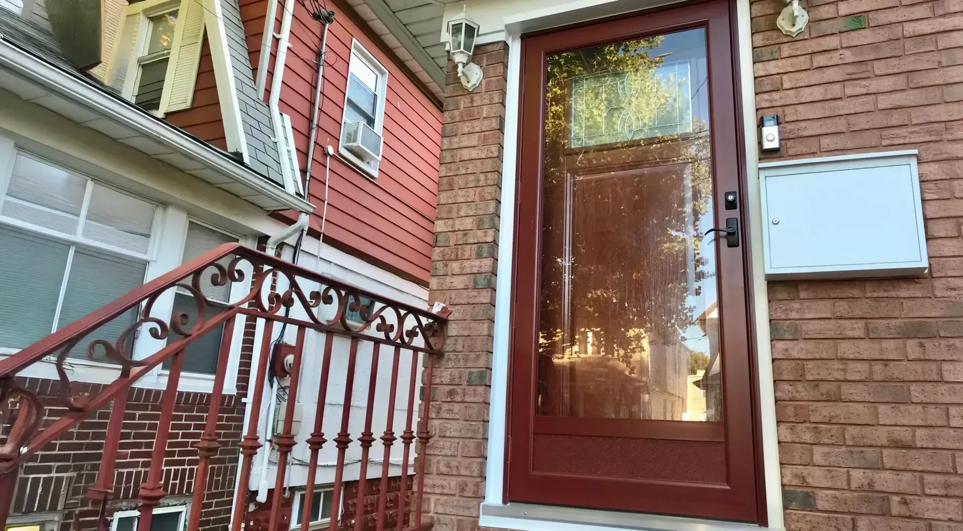 Exterior low-angle view of a dark red storm door with glass panes installed over a main entry on a red brick stoop with a wrought-iron handrail, next to a multi-story home with red siding.
