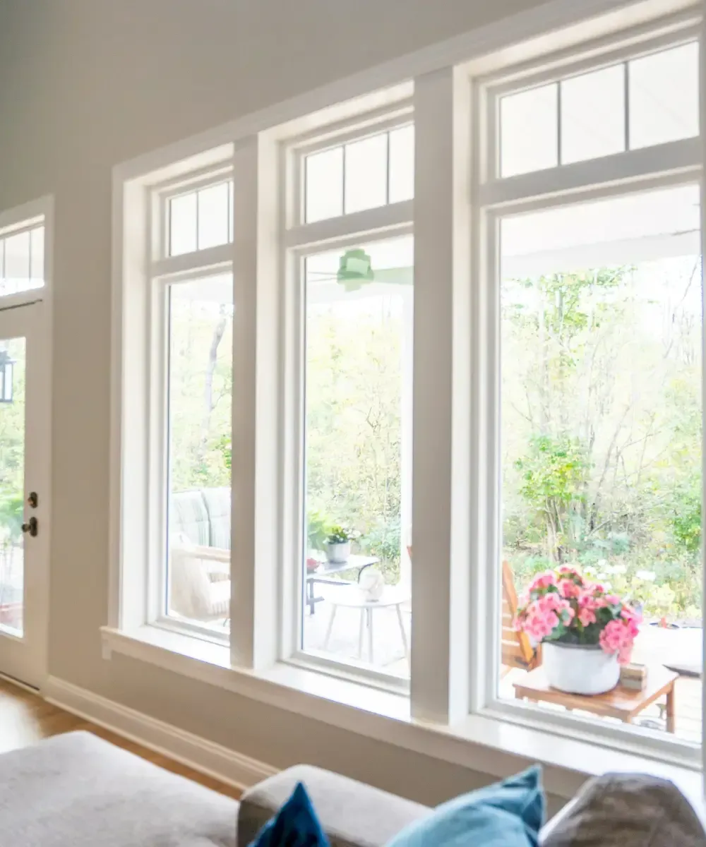 Interior view of a bright room featuring three tall white-framed picture windows topped with slotted transom panes, looking out onto a sunlit patio and garden.