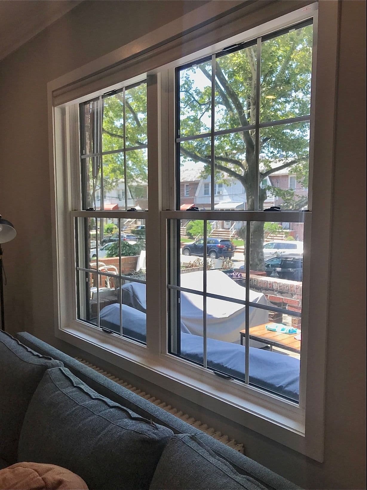 Interior view of a double-hung window set with colonial-style grilles, positioned behind a grey sofa and overlooking a suburban street.
