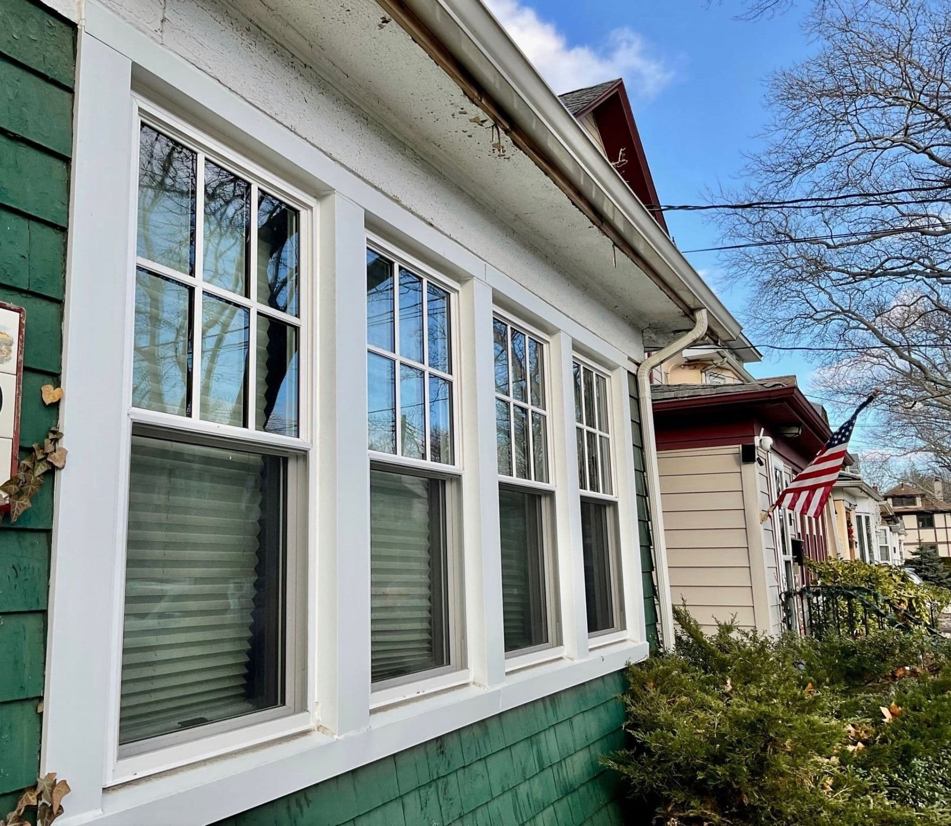 Exterior view of four white double-hung windows with grilles on the upper sashes, set into green shingle siding on a residential house.