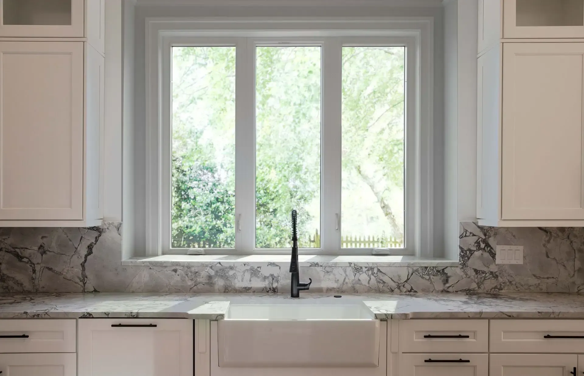 Interior view of a bright kitchen with three white casement windows set above a white farmhouse sink, matte black faucet, and heavily veined marble countertops.