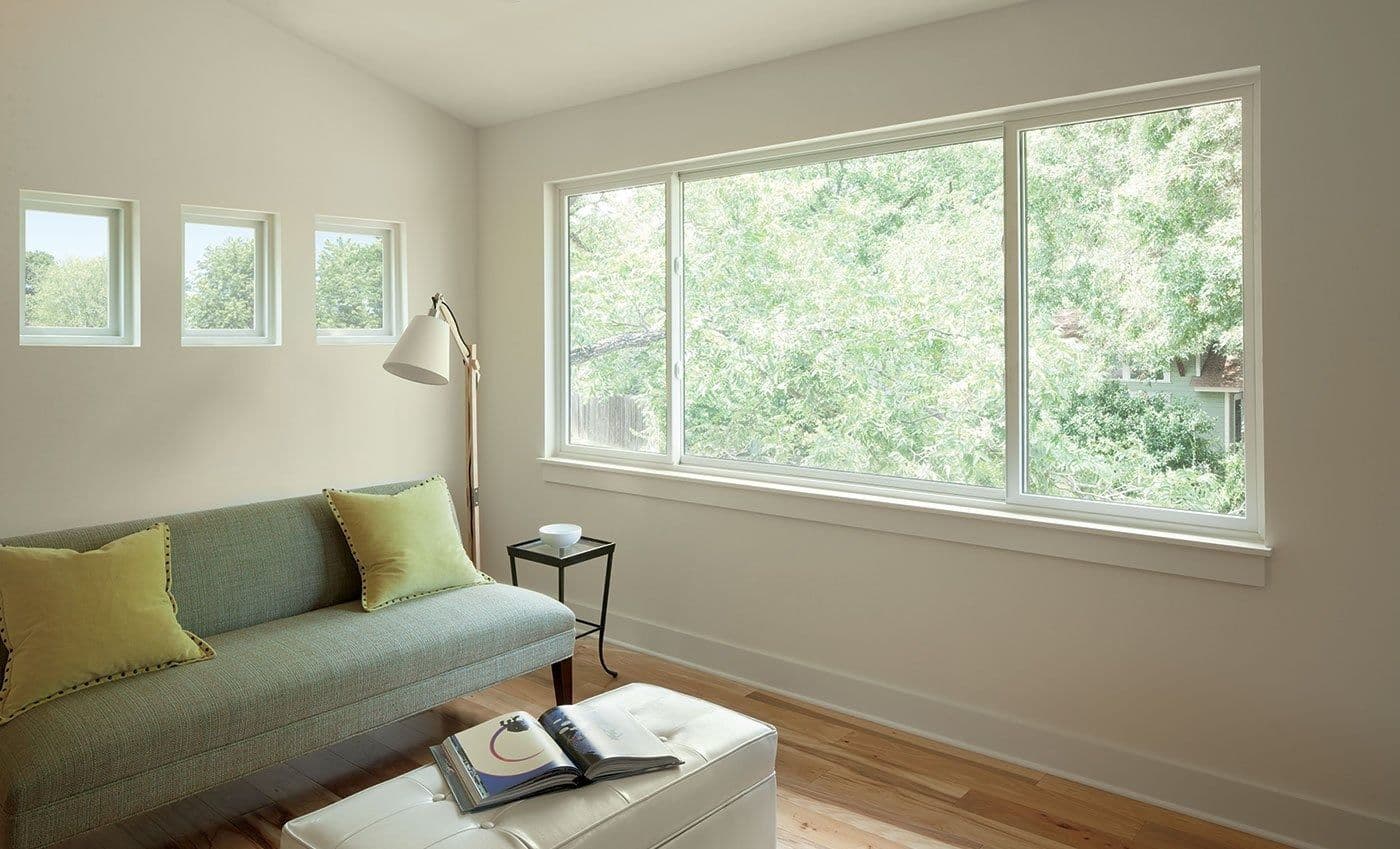 Interior view of a modern, sunlit living room featuring a wide white-framed horizontal sliding window looking out onto lush green tree foliage. Three small square accent windows line the adjacent wall above a sage green sofa with yellow throw pillows, accompanied by a white leather ottoman, a floor lamp, and light wood flooring.