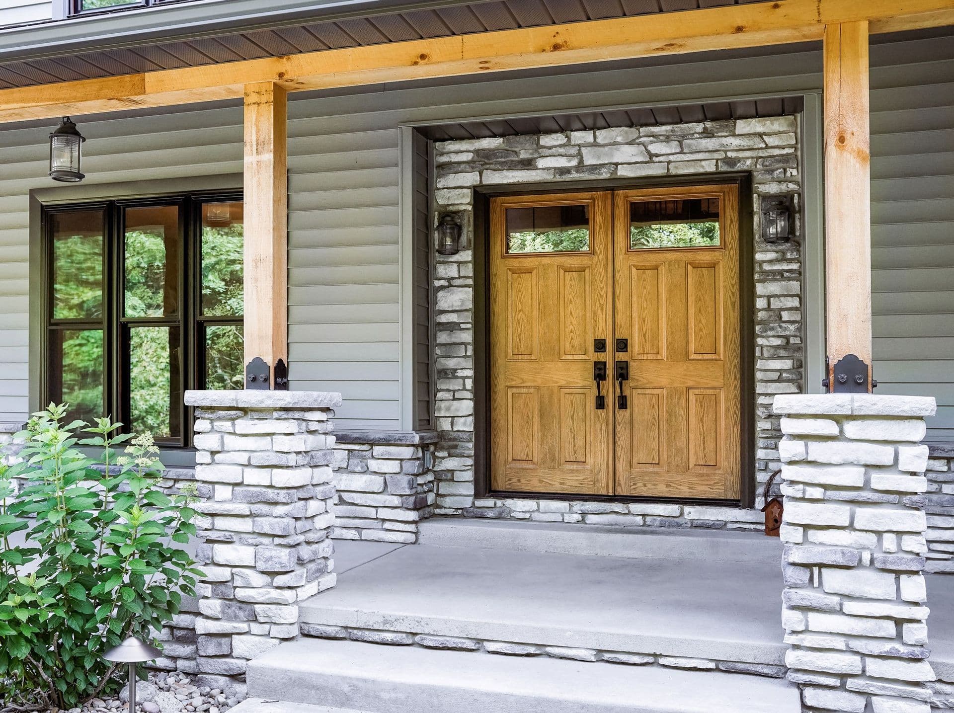 Exterior view of double oak-finished front doors with four raised panels and upper rectangular glass panes, set in a stacked stone entry surround on a covered porch with stone columns and grey horizontal siding.