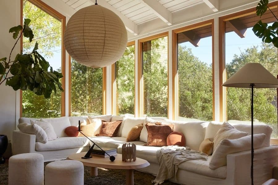 Interior view of a bright sunroom with tall, floor-to-ceiling wood-framed picture windows wrapping around a white sectional sofa, highlighted by a large paper lantern pendant light and leafy indoor houseplants.