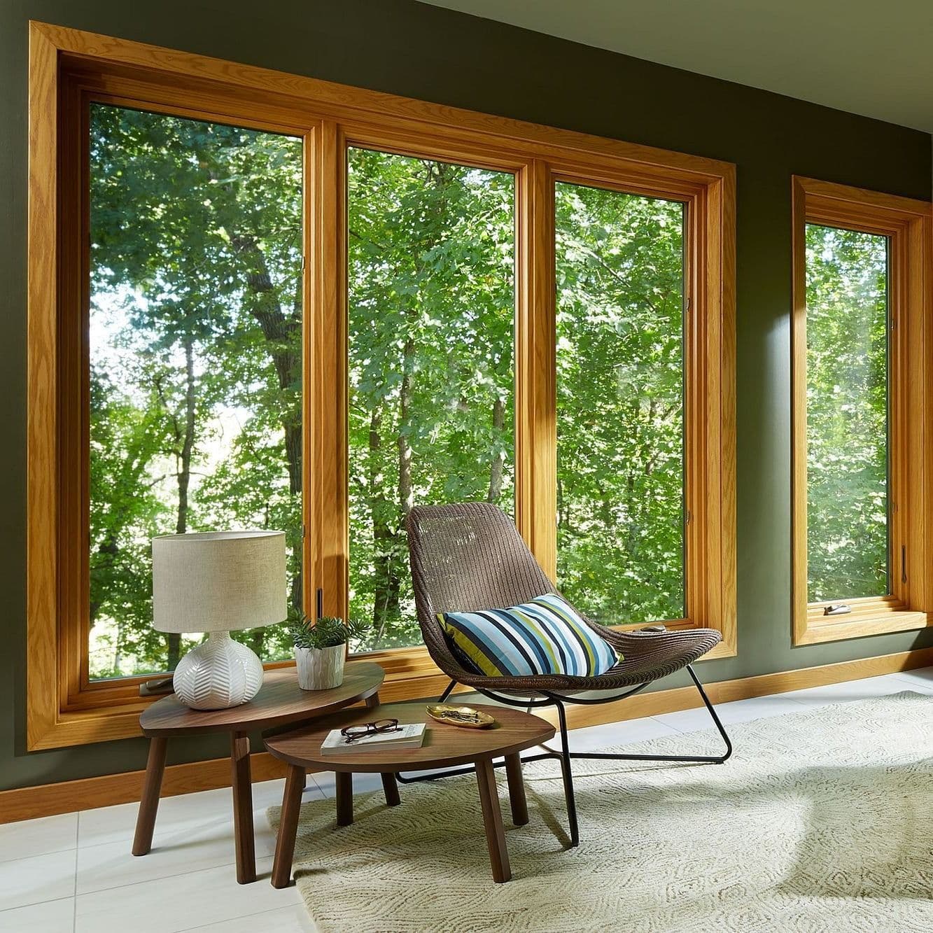 Tall wood-grain interior casement windows overlooking a lush green forest, featured in a room with olive green walls, a woven lounge chair, nested coffee tables, and a table lamp.