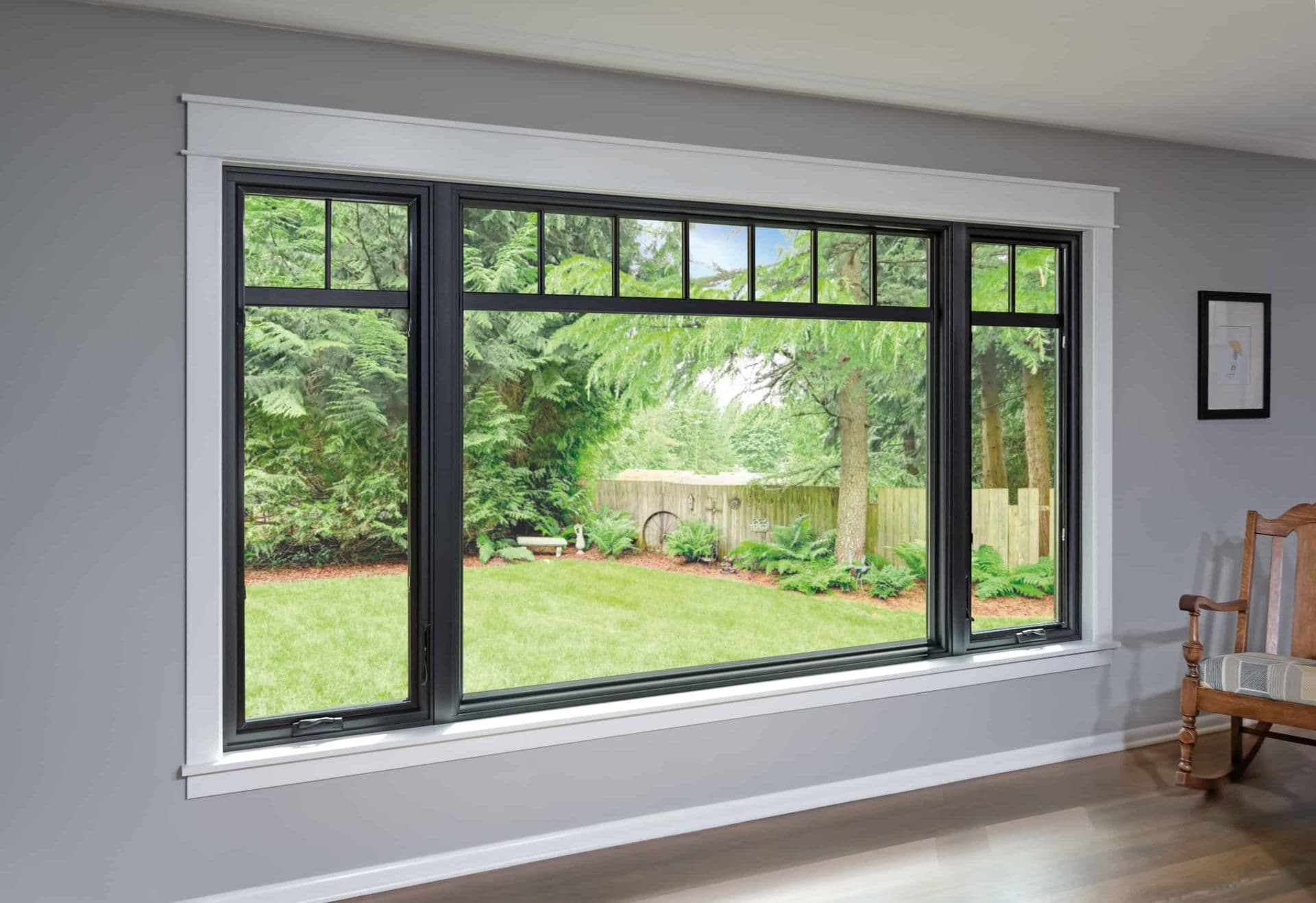 Interior view of a large black-framed picture window set flanked by operable casement windows with continuous upper grilles, encased in white molding on a grey wall and looking out onto a grassy backyard.