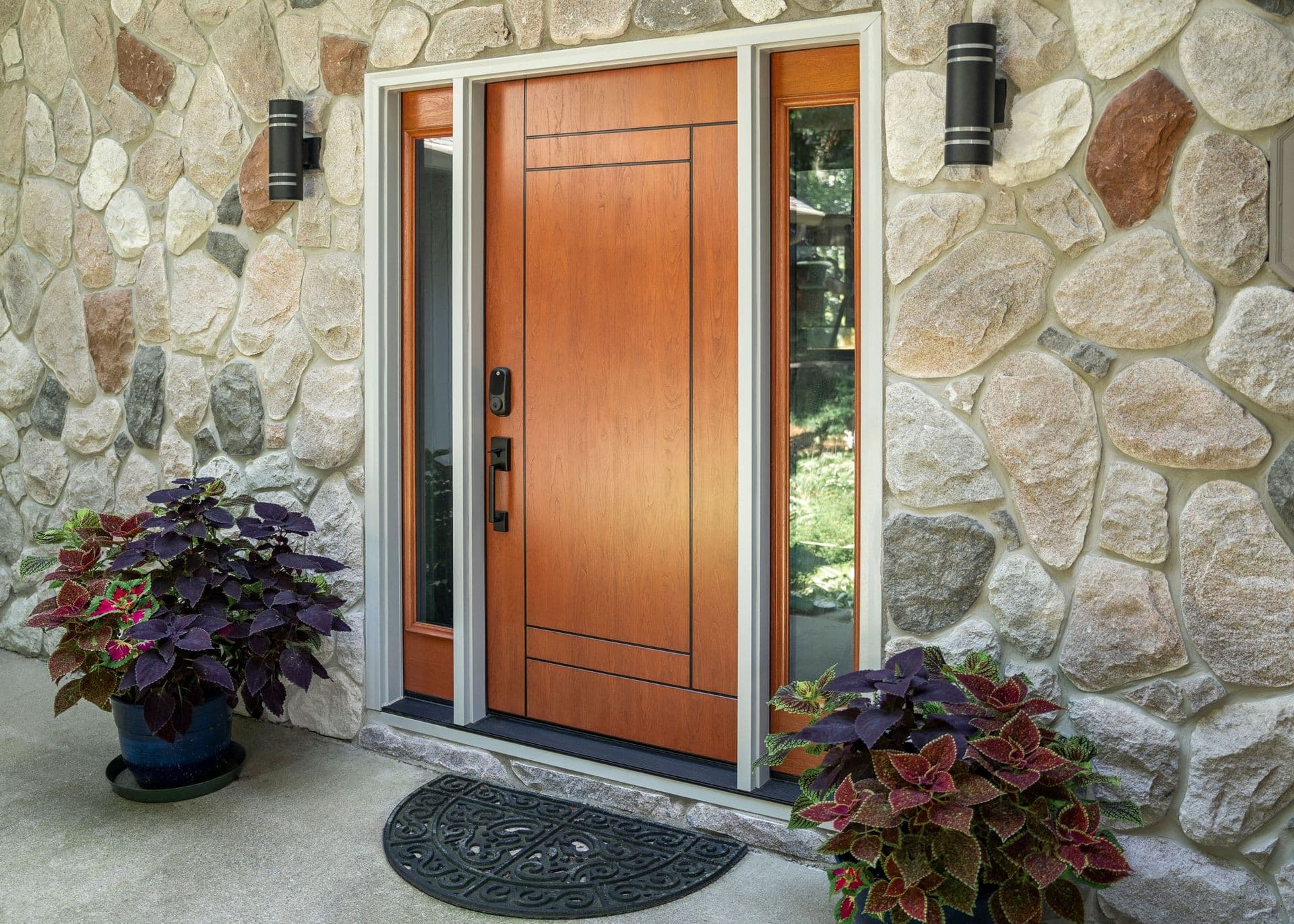Exterior view of a modern toffee-stained wood front door featuring clean linear inscribed geometric panel details, flanked by two full-length clear glass sidelights, installed into a light-colored fieldstone wall.