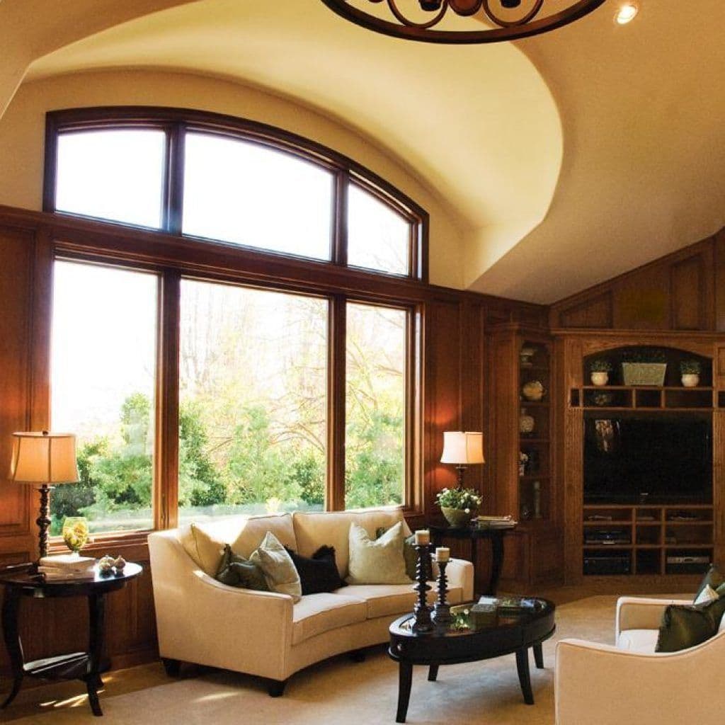 Large wood-framed window featuring a prominent arched upper transom, set in a cozy, wood-paneled living room with a beige sofa, dark coffee table, and custom built-in cabinetry.
