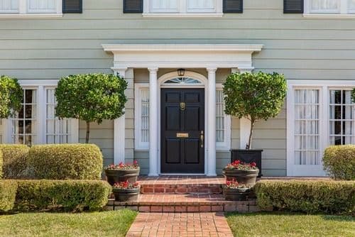 Traditional home entrance with a black front door, white columns, red brick walkway, trimmed hedges, and potted flowers.