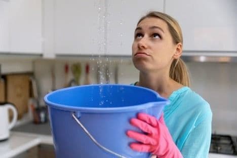 Woman holding a blue bucket beneath water leaking from the ceiling in a kitchen.