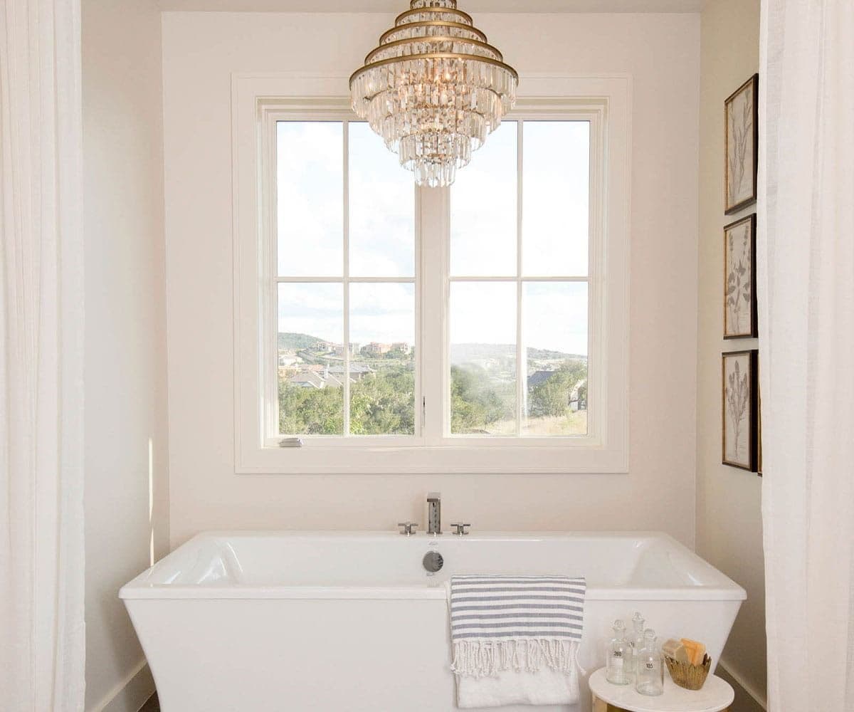 Bright bathroom with a white freestanding bathtub beneath a large window and crystal chandelier.