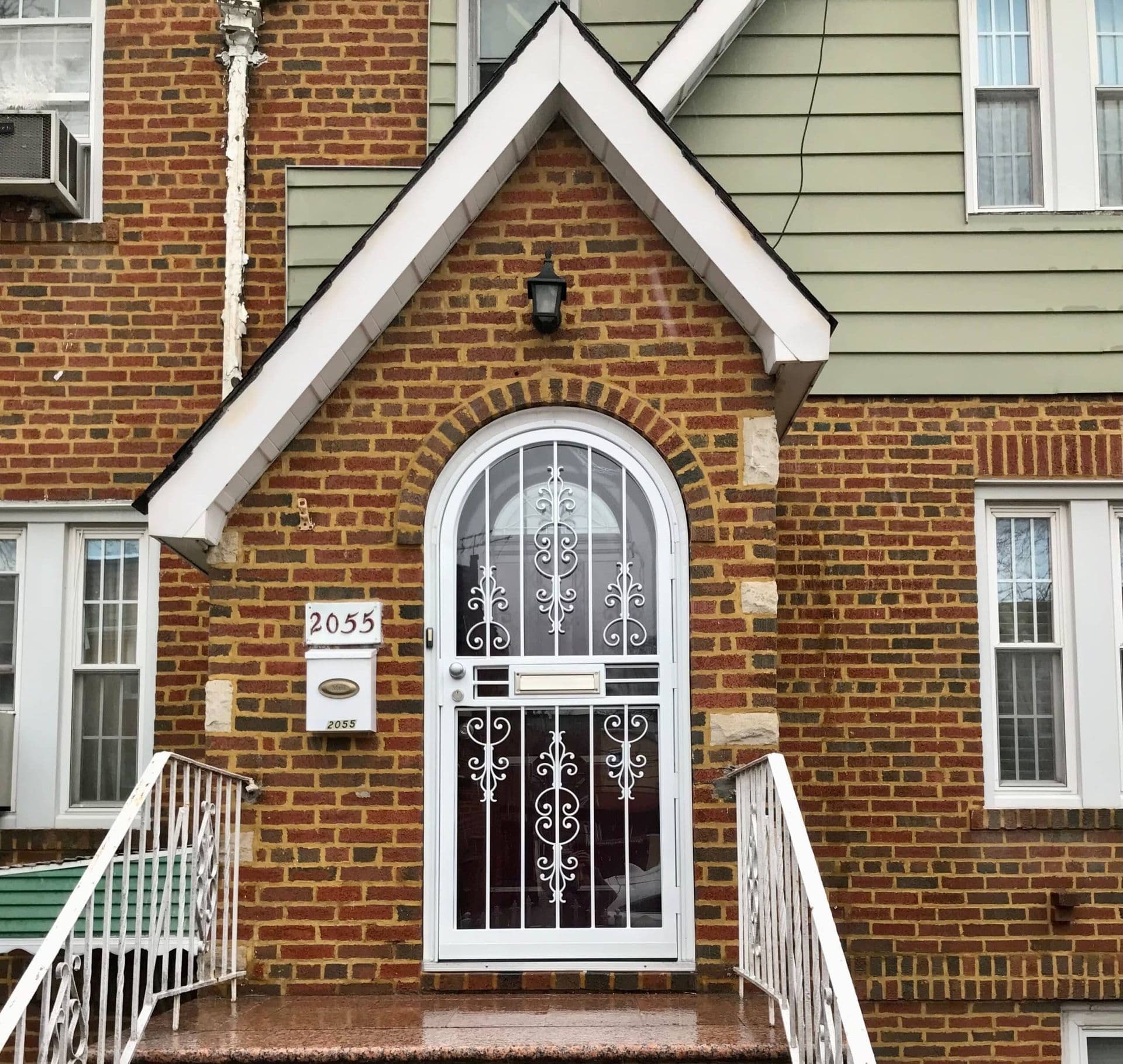White arched security door with decorative metal scrollwork installed at the entrance of a red brick home.