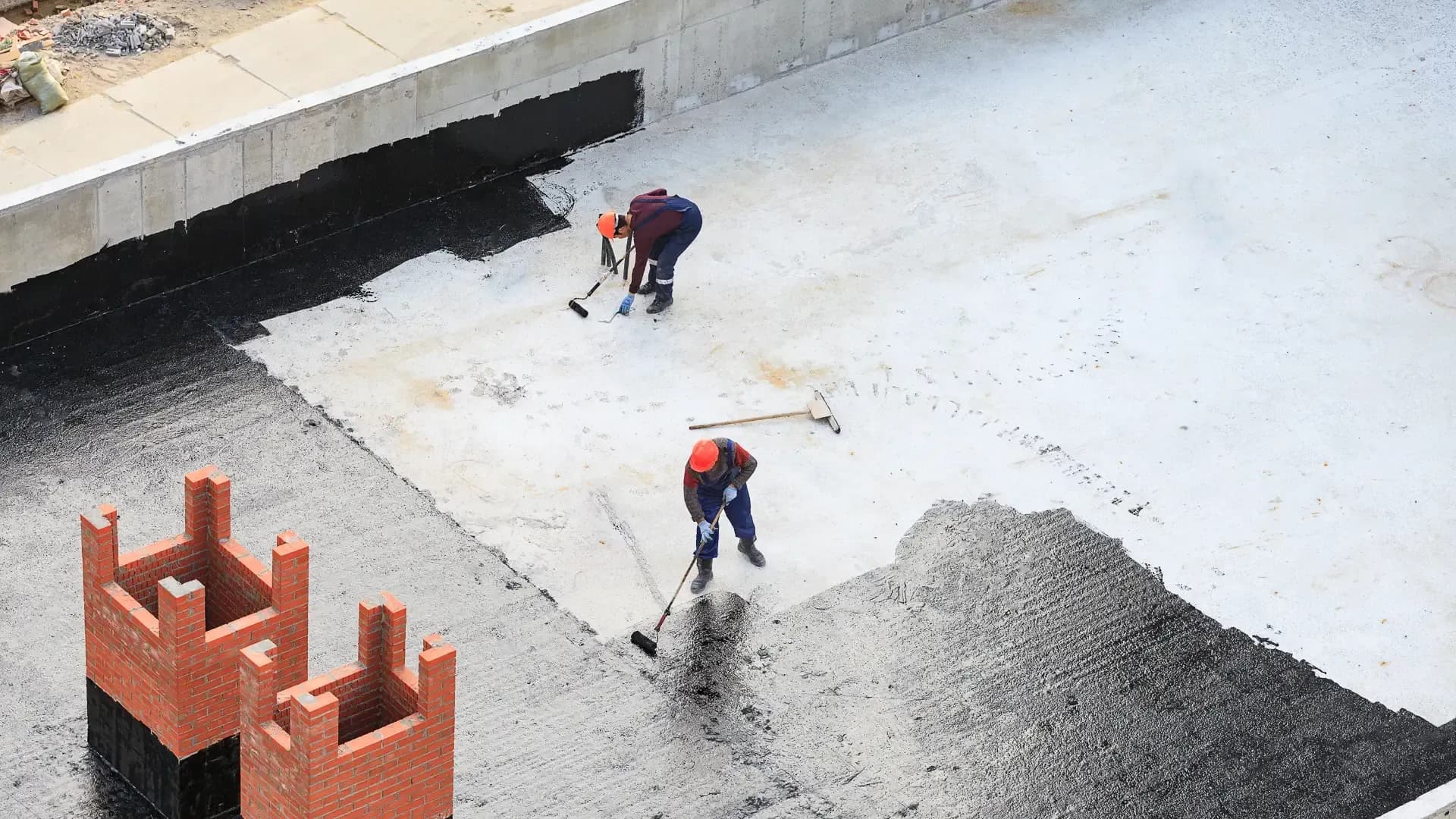 Two construction workers applying black waterproofing material to a concrete foundation and surrounding walls.
