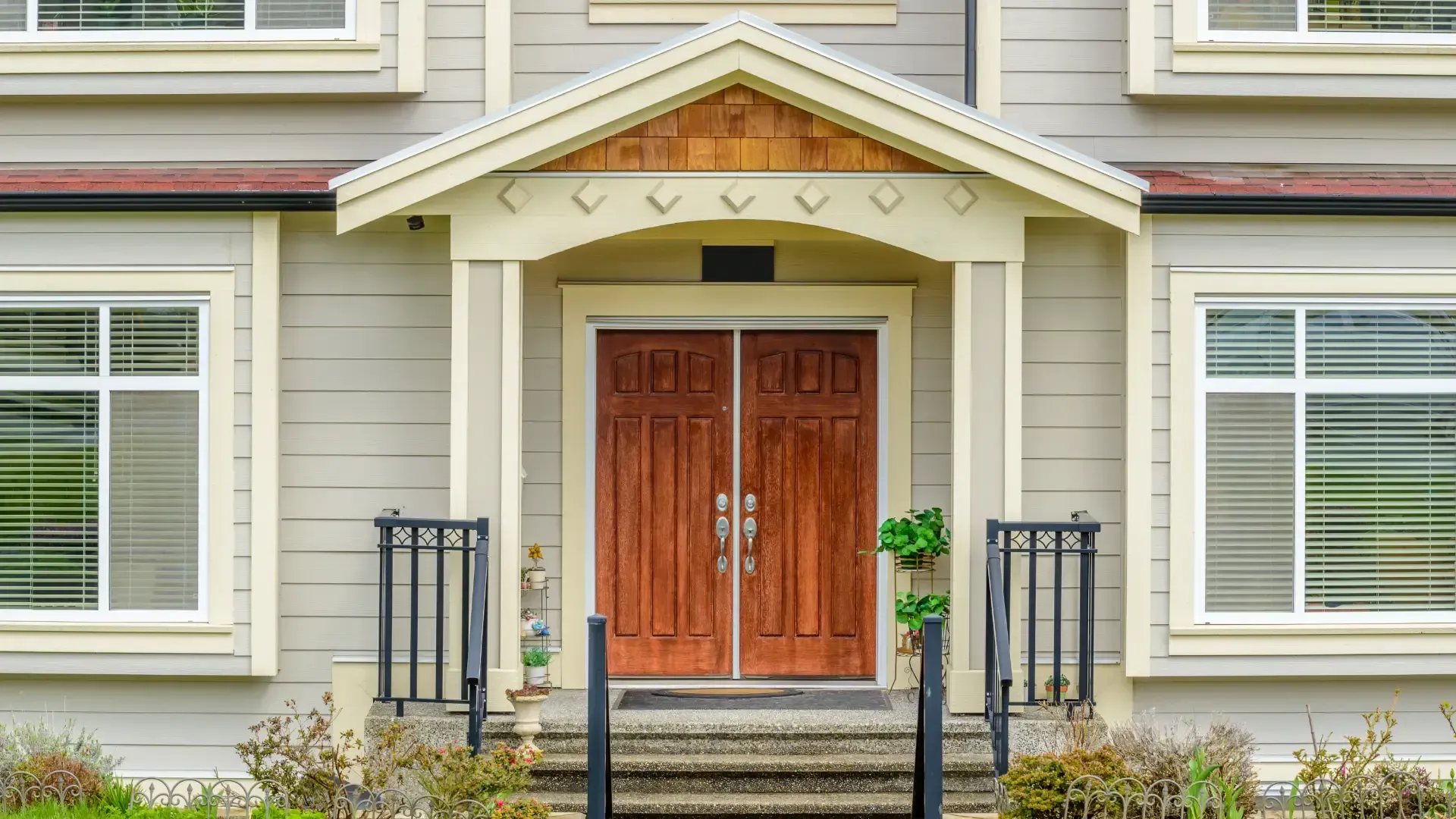 Covered front entrance with double wooden doors, beige siding, white-trimmed windows, black railings, and landscaped steps.