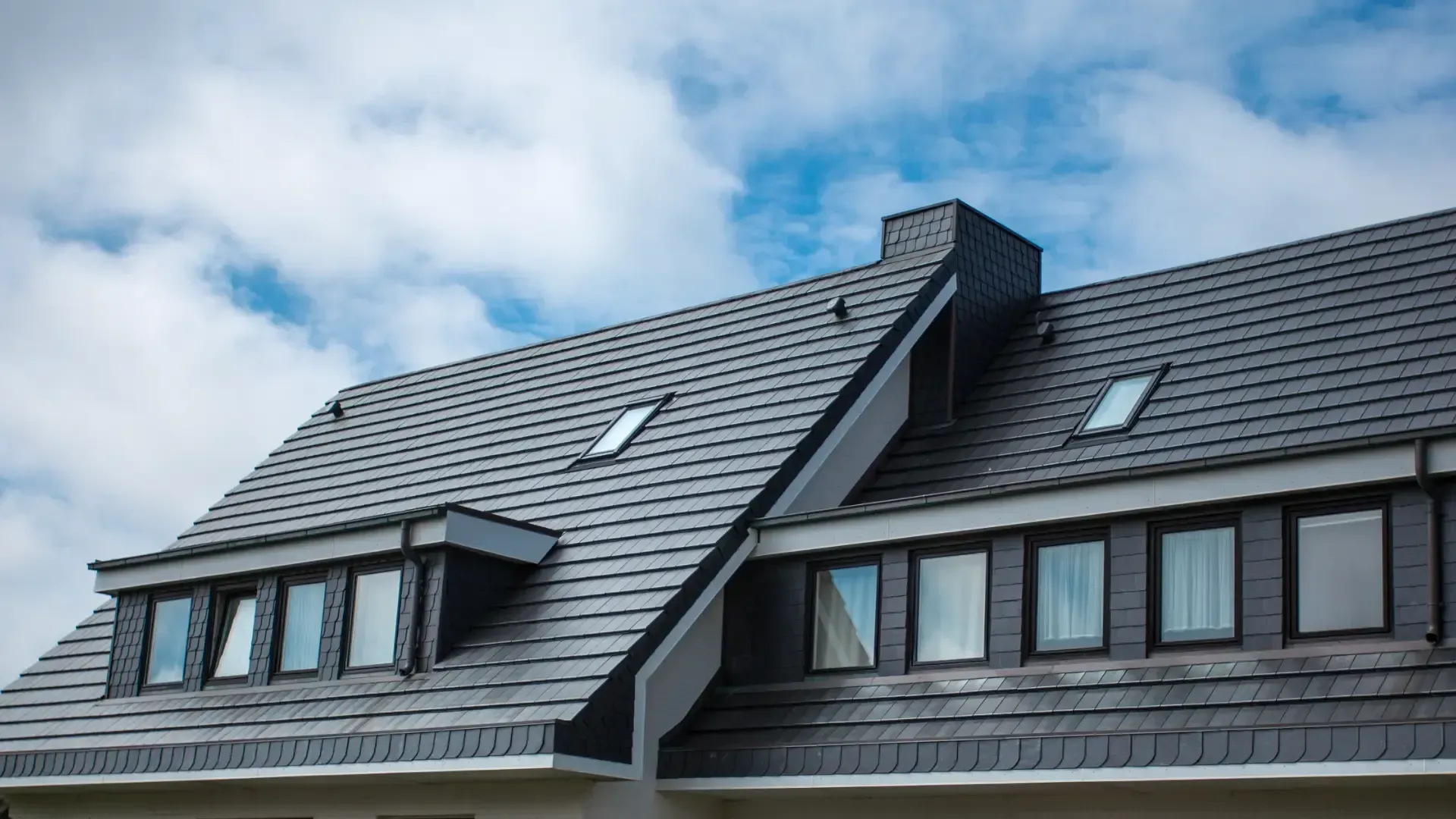 Dark gray tiled roof with skylights, dormer windows, and a matching chimney beneath a partly cloudy sky.