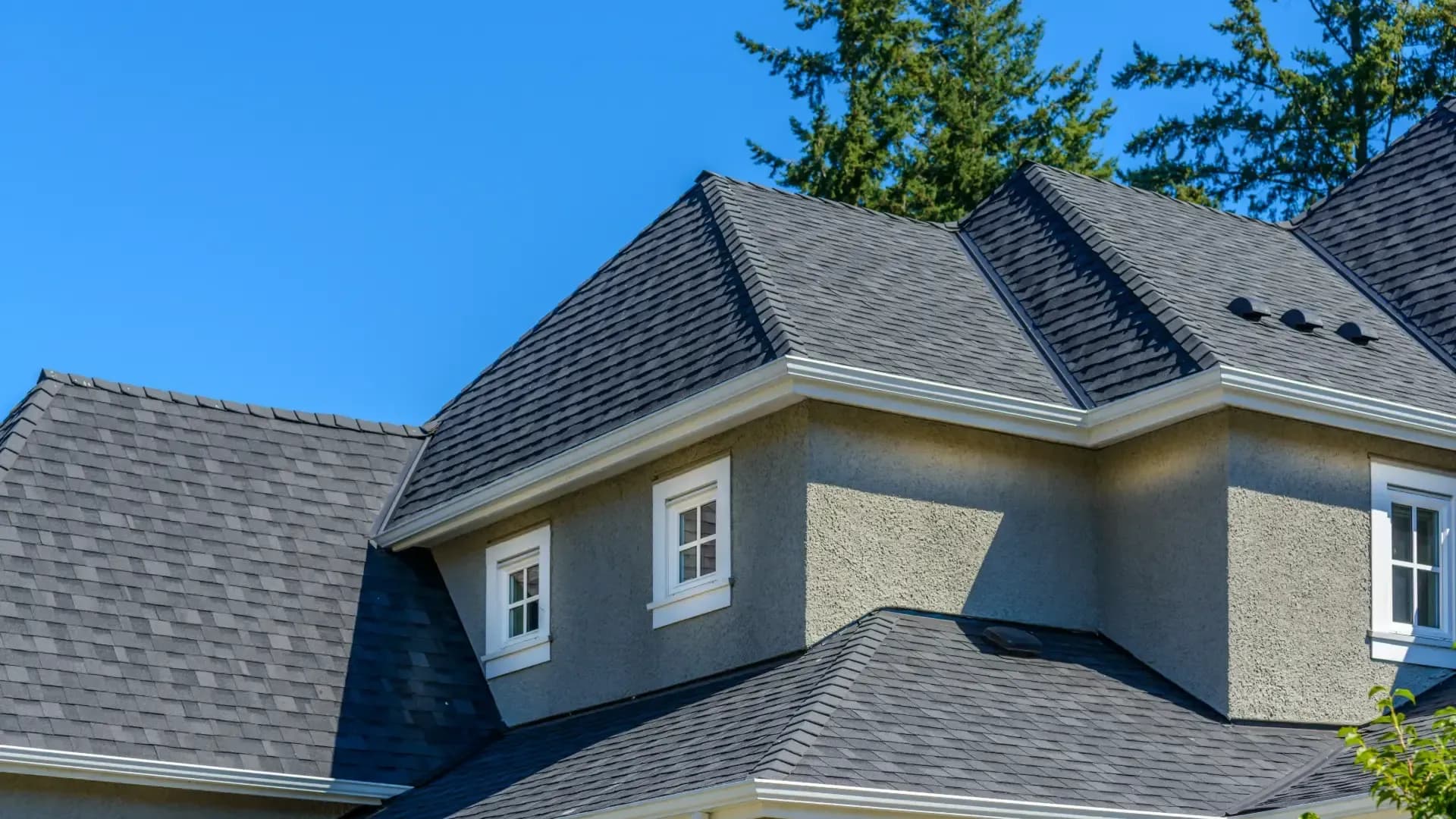 Large home with multiple dark gray asphalt shingle roof sections, white gutters, gray stucco, and white-trimmed windows.
