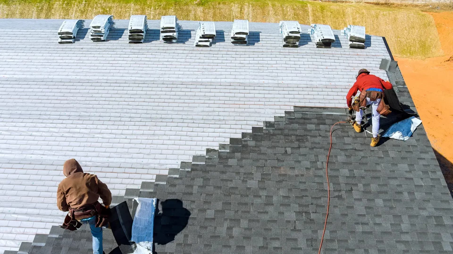 Two roofing contractors installing dark asphalt shingles on a large sloped roof with bundles of roofing materials nearby.