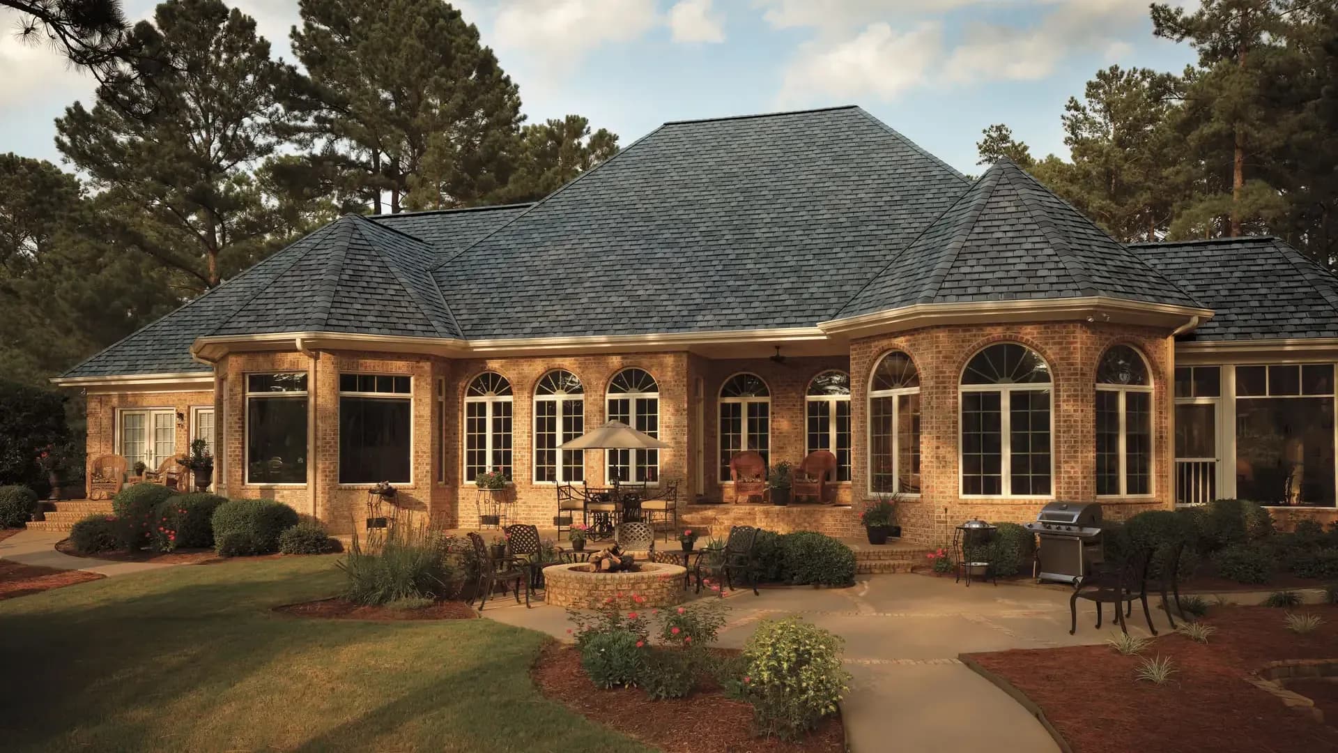 Wide exterior view of a sprawling red brick estate featuring arched windows, a dark grey antique slate-style shingle roof, and a paved backyard patio equipped with an outdoor fire pit and seating.