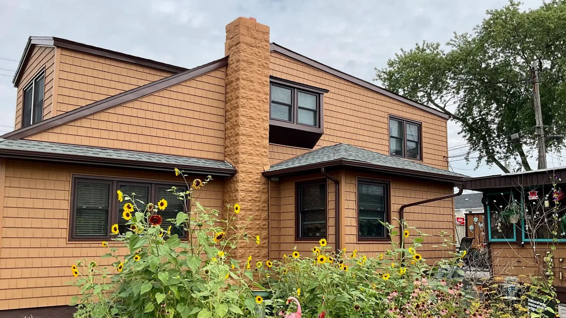 Two-story home with warm brown shake siding, dark-trimmed windows, a stone chimney, and sunflowers in the garden.