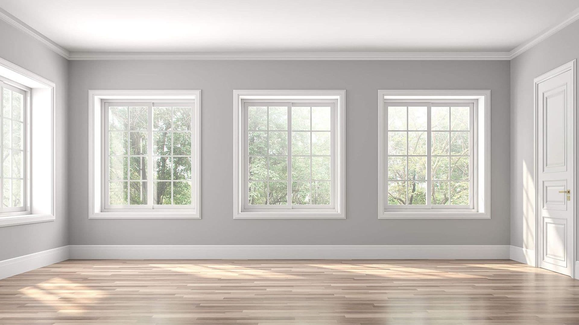 Bright empty room with gray walls, wood flooring, white trim, and four large vinyl windows overlooking trees.