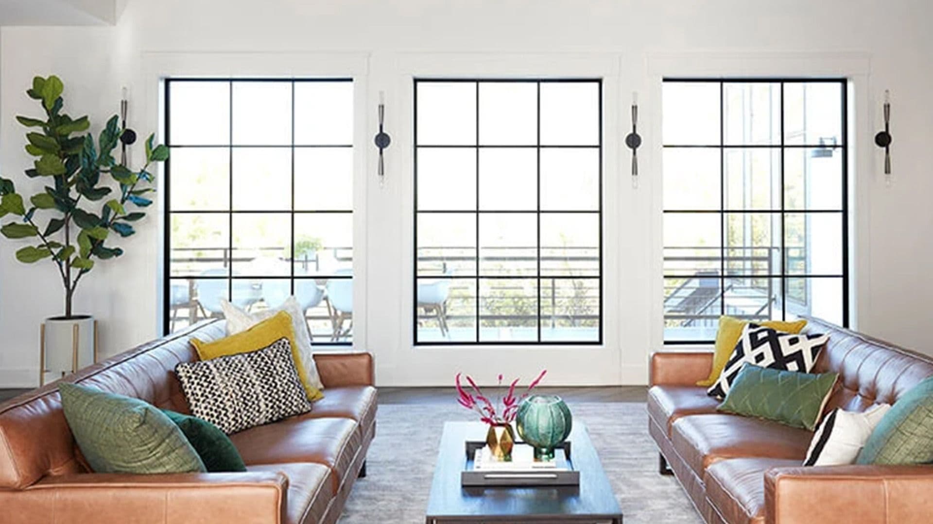 Modern living room with brown leather sofas and three large black-grid windows overlooking a patio.