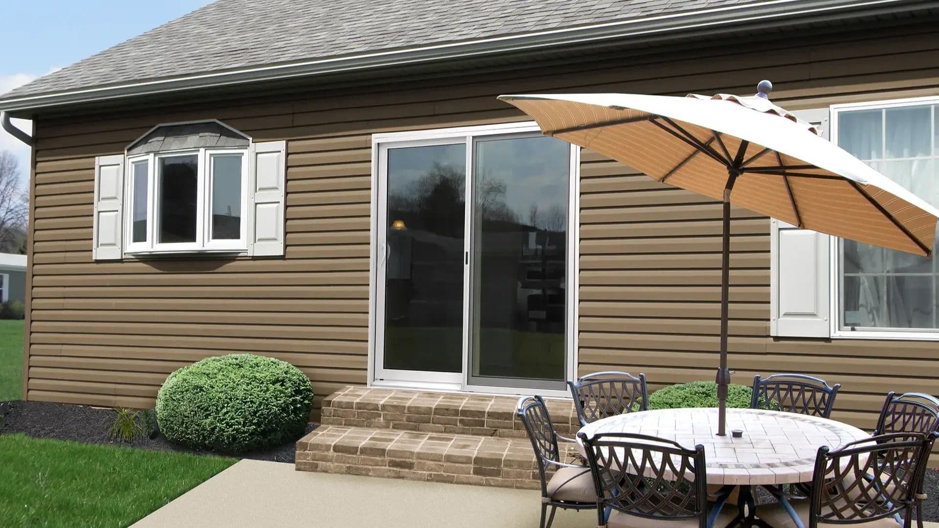 Backyard patio with a white sliding glass door, bay window, brown siding, outdoor dining table, and umbrella.